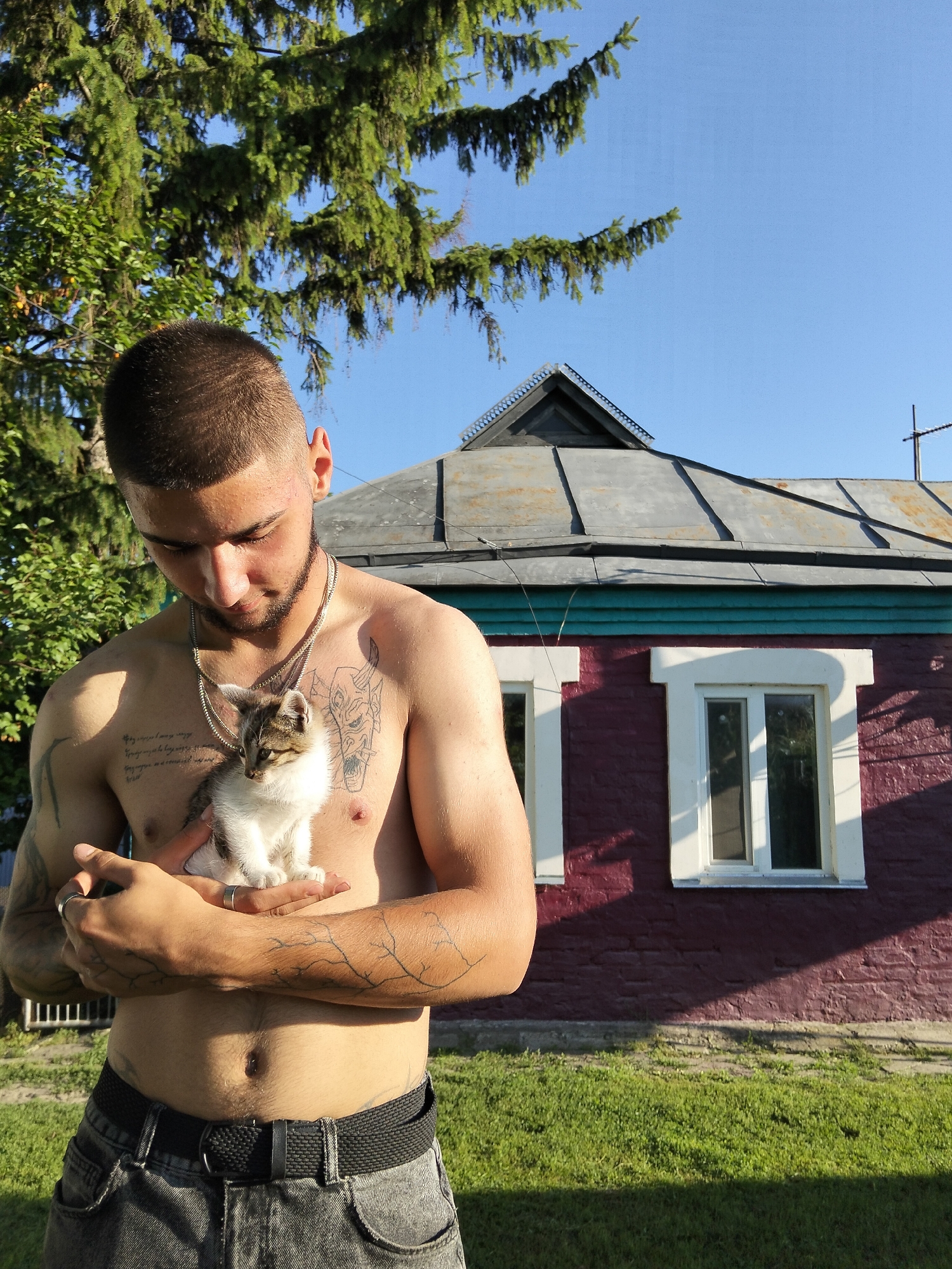 A young man with a short buzz cut and a beard stands in front of a maroon-colored house in Kliushnykivka, Ukraine. He is shirtless, revealing tattoos on his chest and arms, and is wearing dark jeans and a black belt. The man is holding a small tabby and white kitten in his hands, cradling it against his chest. The kitten is looking to the side. The house has a grey, corrugated metal roof and two white-trimmed windows. The background shows green trees and a clear blue sky. The scene is lit by bright sunlight, casting shadows on the house and ground.