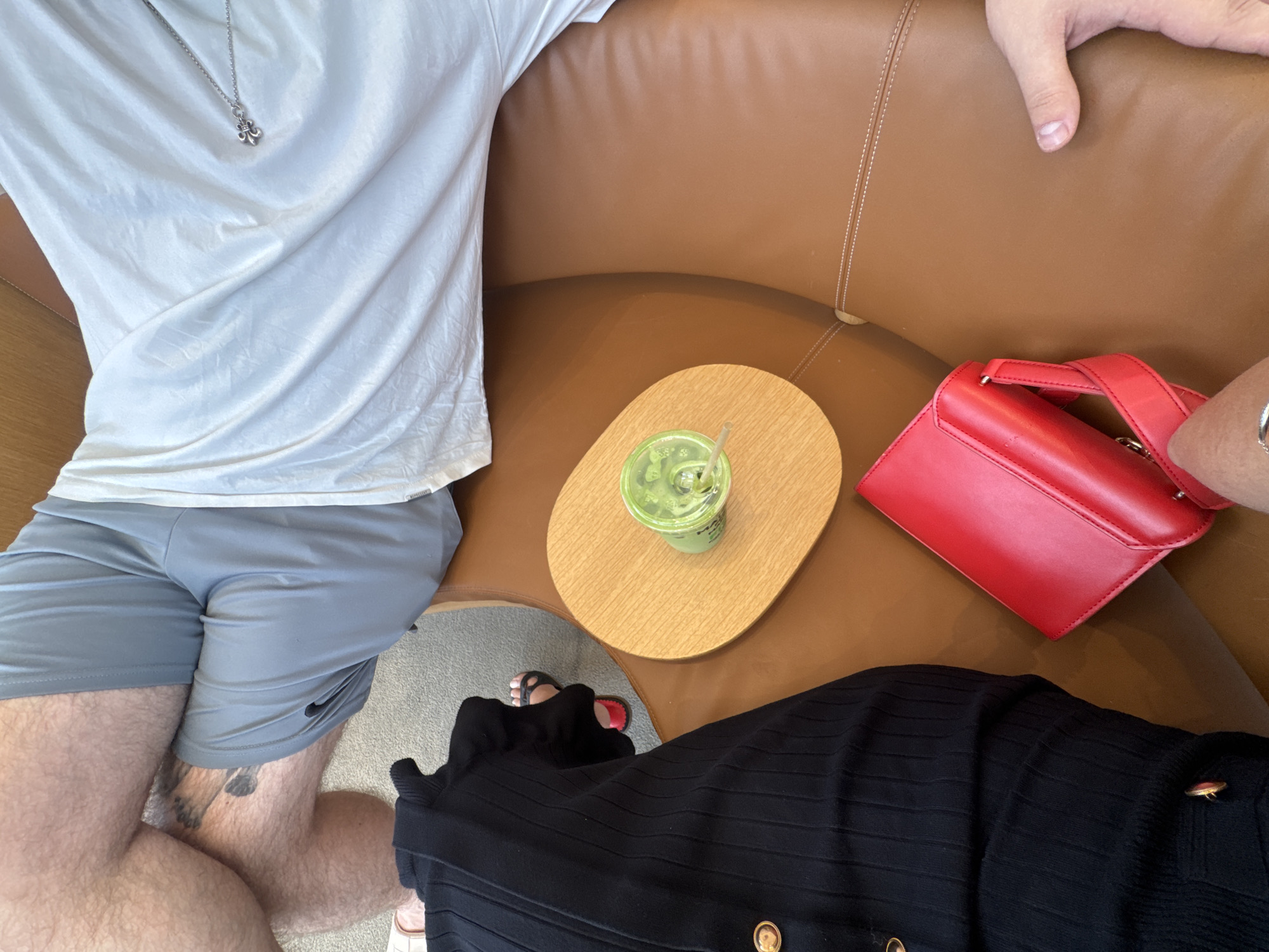 In Aventura, Florida, USA, two individuals are seated on a brown leather couch. One person is wearing grey shorts and a white t-shirt, with a silver necklace visible. The other person is dressed in a black ribbed top and is wearing black sandals with red accents. Between them rests a wooden coaster topped with a clear cup of matcha green tea and a straw. To the right of the cup, a bright red clutch purse is placed on the couch. The setting appears to be a comfortable indoor environment, possibly a lounge or living room.