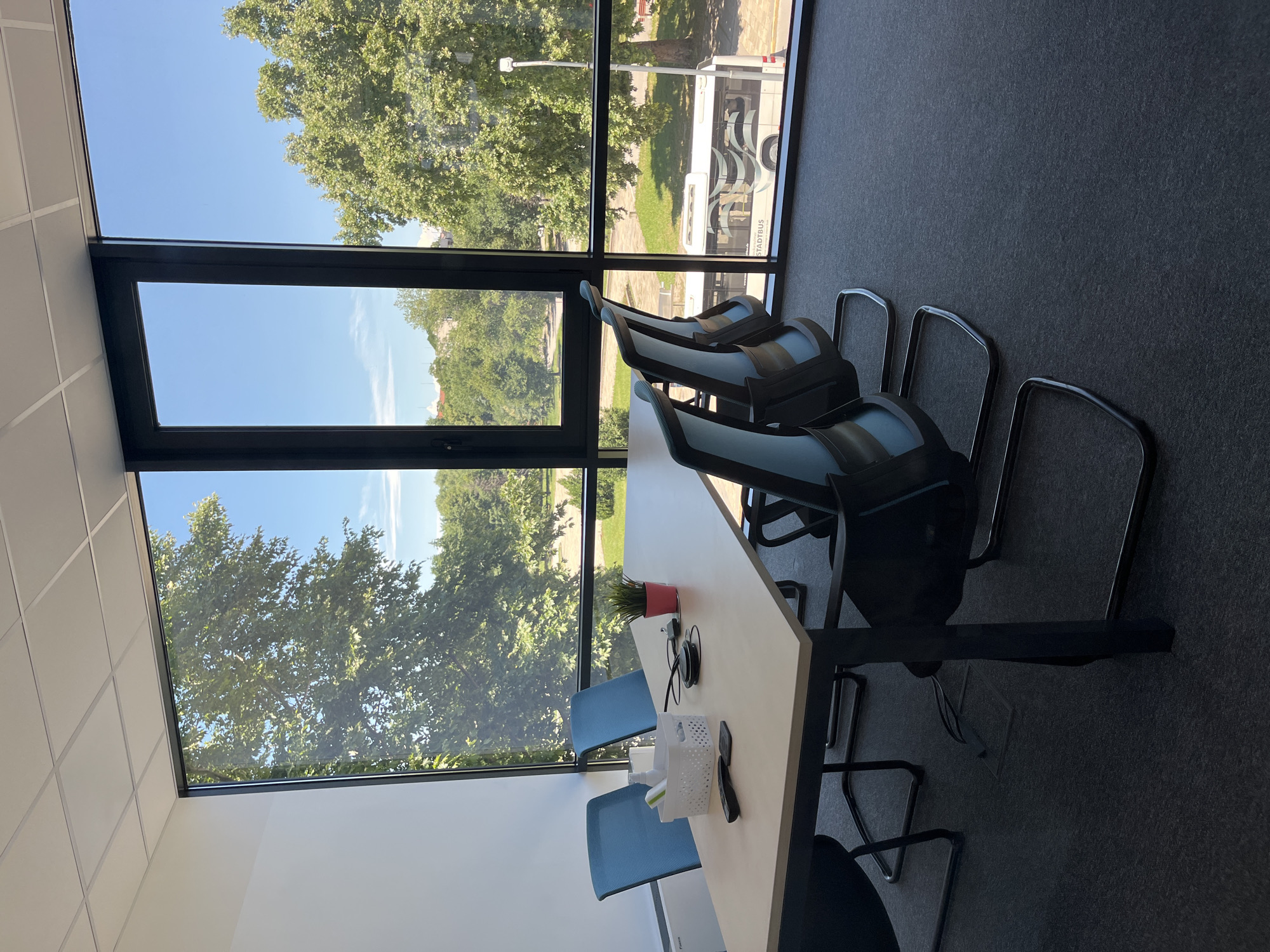 This is a forensic, matter-of-fact description of a meeting room with a view of trees and a street in Plovdiv, Bulgaria. The room has a rectangular table with four office chairs around it. The chairs are black with light blue upholstery. On the table, there is a white basket containing a bottle of hand sanitizer and other items, a pink pot with a small green plant, and two black remote controls. Cables are visible on the table, connecting to conference call equipment. Through the large windows, a park-like setting with mature trees and a clear blue sky is visible. In the background, a white bus with "STADTBUS" written on it is parked on the street. The floor of the room is carpeted in a dark grey material. The room appears to be empty of people.