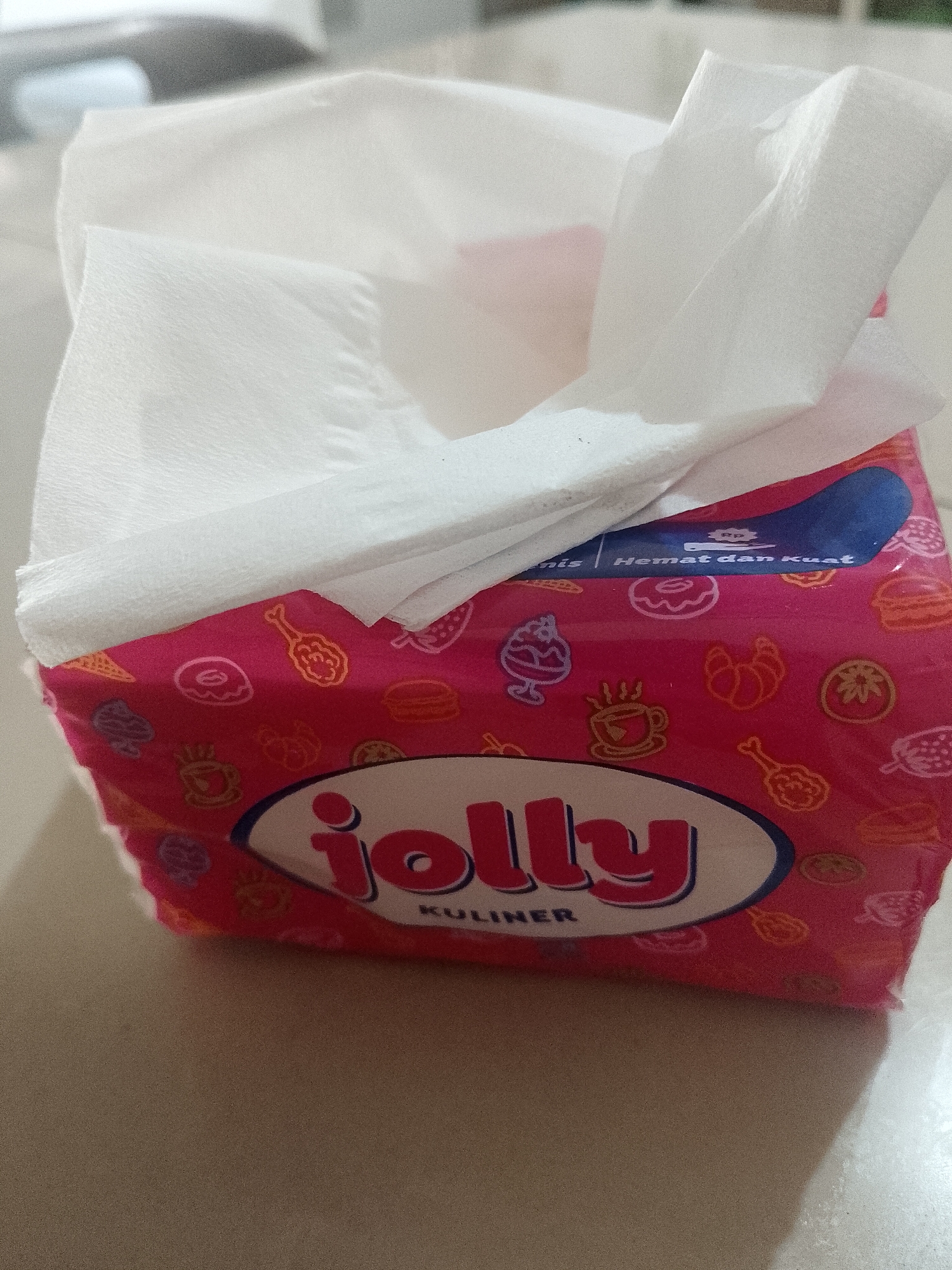 The image shows a pack of "Jolly Kuliner" tissues on a light-colored surface. The tissue box is pink with a repeating pattern of food items, and the brand name "Jolly" is prominently displayed in a white oval with red lettering. Some tissues are pulled up and out of the box. The location in Lhokseumawe, Indonesia, is not evident in the provided image.