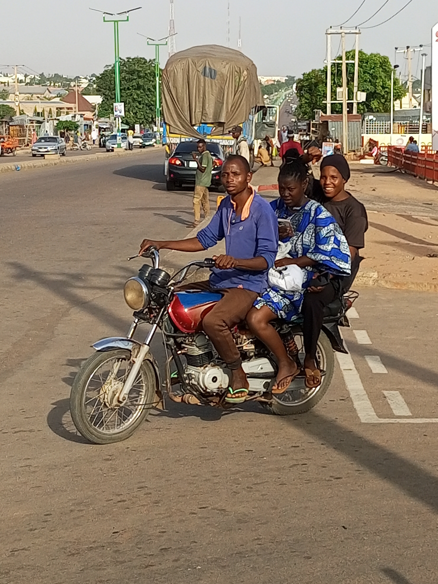 In Tempure, Nigeria, a man rides a motorcycle with three passengers on a paved road. The rider, wearing a blue shirt and brown pants, controls the motorcycle. Behind him, a woman in a blue and white patterned dress holds a white bag, and another woman in a black headscarf and black top is seated behind her. All three passengers are positioned behind the rider on the motorcycle's seat. The motorcycle has a red and blue fuel tank and chrome accents. In the background, a large truck covered with a tarp is visible, along with other vehicles, buildings, and streetlights, indicating an urban environment. The road has white painted lane markings. The lighting suggests it is daytime.