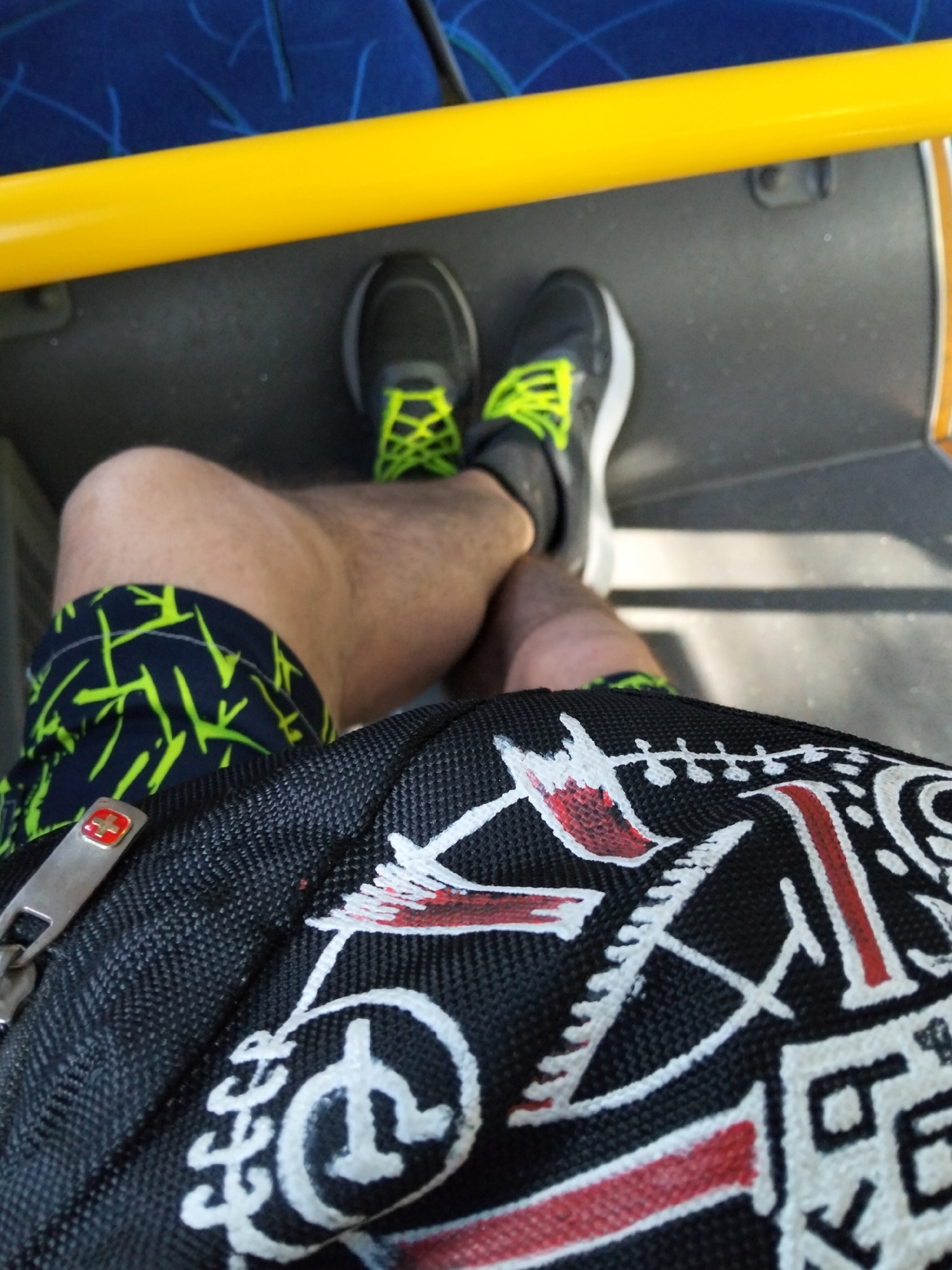 The image shows a close-up view of a person's legs and a bag, likely inside a public transport vehicle. The person is wearing black and neon green athletic shoes and dark shorts with a neon green geometric pattern. A black bag with white and red graphic designs is in the foreground, with a silver zipper pull featuring a red and white emblem. The background shows a yellow railing and blue seating, characteristic of a bus or tram interior. The scene is dimly lit, suggesting it might be indoors or during cloudy weather. There is no specific indication of the location within Almaty, Kazakhstan, as the elements are generic to public transportation.