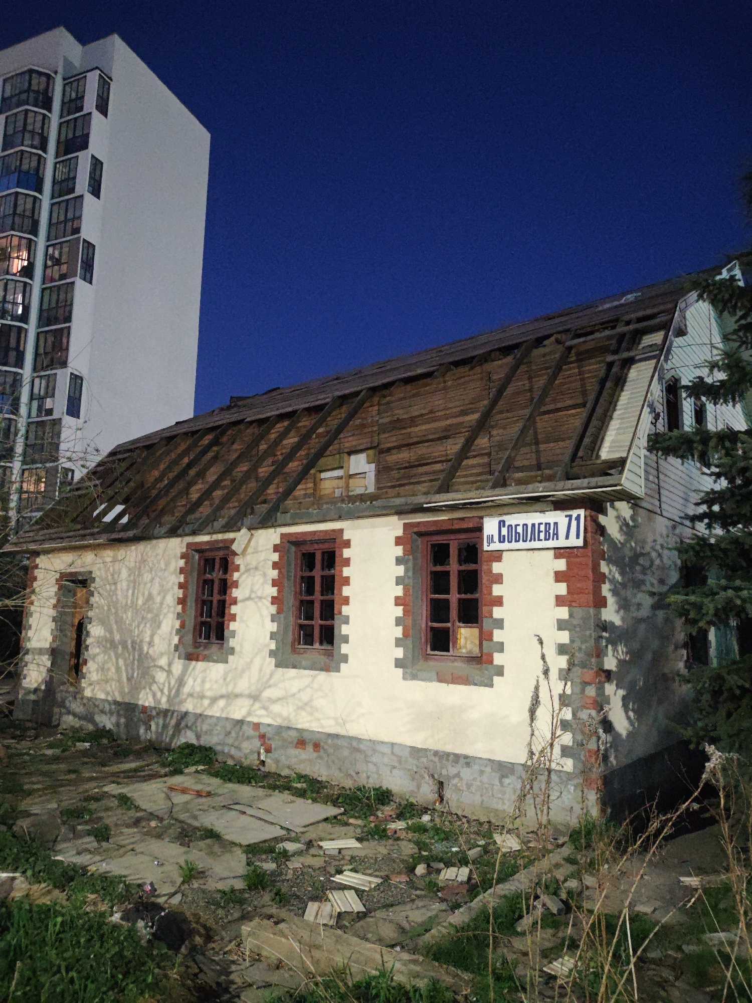 The image depicts a dilapidated two-story building in Yekaterinburg, Russia, identifiable by a street sign reading "ul. Soboleva 71" (Soboleva Street 71). The structure's roof is largely missing, exposing the wooden frame. The building's facade is painted white with brick trim around the windows and base. The windows are dark and appear to be boarded up or broken. The surrounding ground is covered in debris, broken concrete slabs, and sparse vegetation. To the left of the building, a modern high-rise apartment building rises sharply. The time of day appears to be night, as indicated by the dark blue sky.