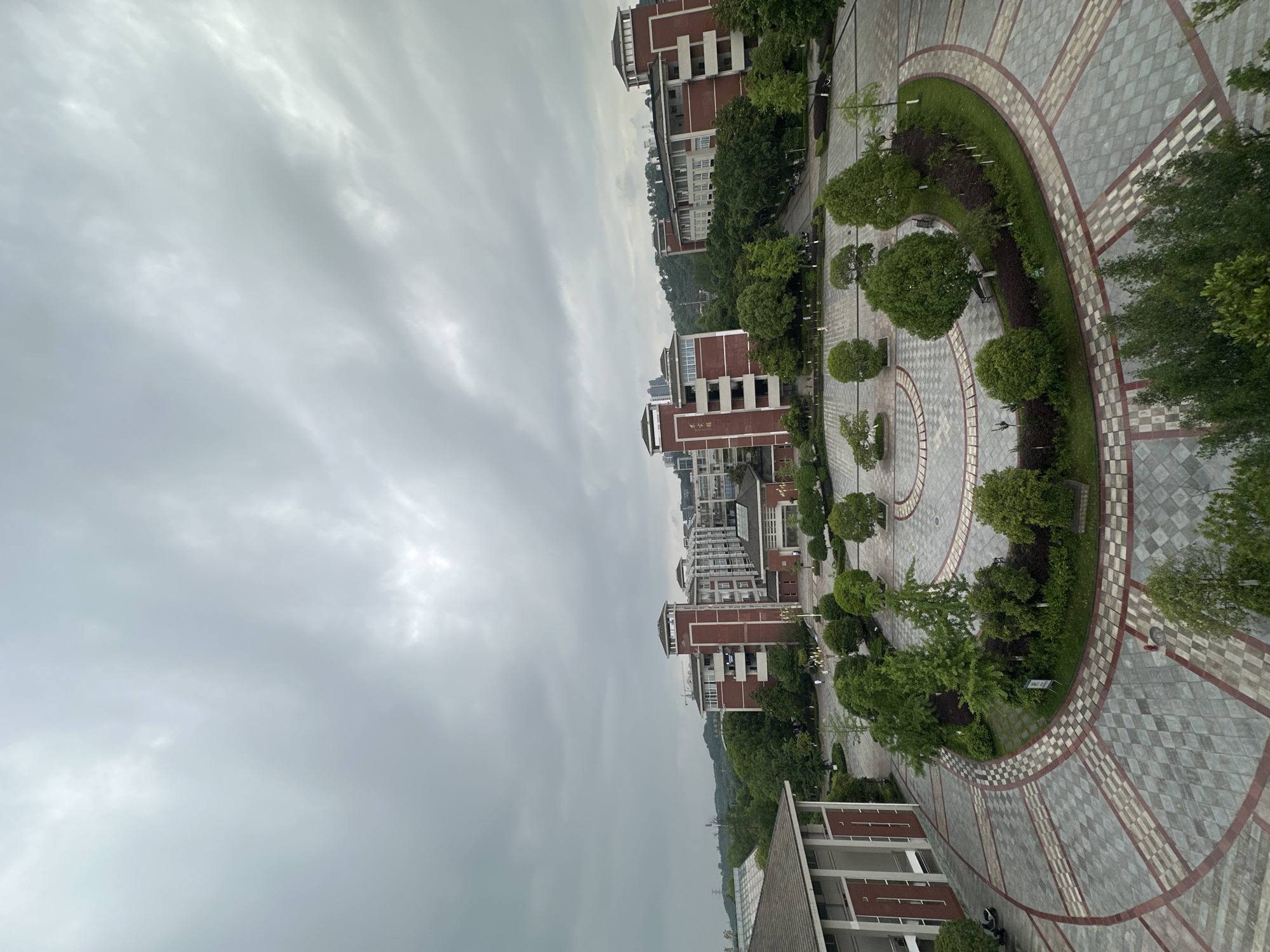 The image shows an elevated view of a campus or university setting in Chongqing High-tech Zone, China. The sky is overcast with striated cloud formations. Several multi-story brick buildings with light-colored accents line the scene from left to right. The buildings appear to be of similar architectural style. A paved plaza featuring a circular design with concentric rings of patterned tiles and landscaped areas with trees and shrubs dominates the foreground. There are no visible people. The overall environment suggests a formal or institutional setting, possibly a university or college campus.