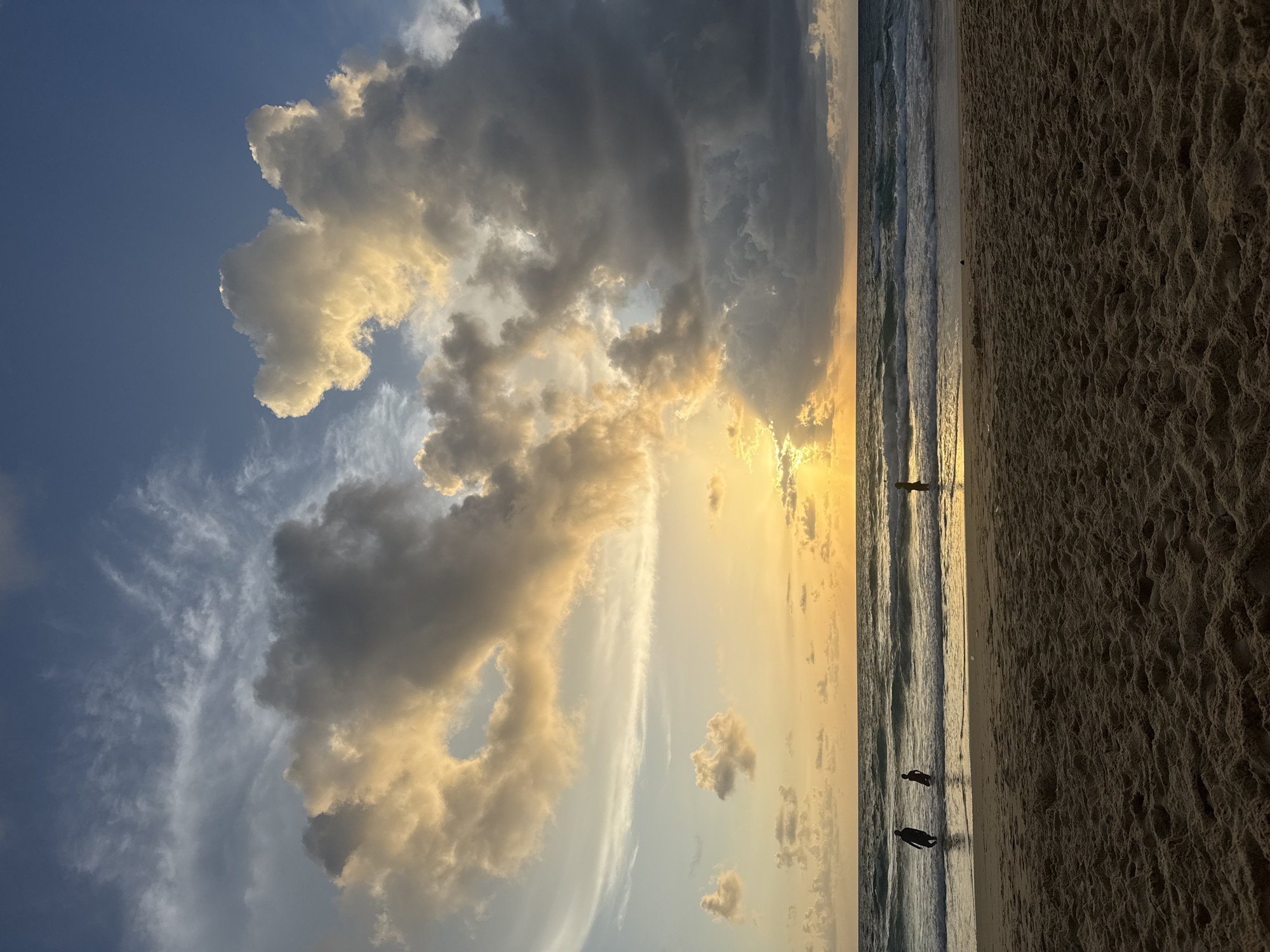 This is a vertical, eye-level, full-shot photograph of Karon Beach in Karon, Thailand, at sunset. The foreground is dominated by the textured, rippled sand of the beach. The middle ground features the ocean with gentle waves rolling onto the shore. In the water, three figures are visible: two are silhouetted against the water and appear to be standing or wading, while the third, further to the right, is standing in the water. The horizon line is clearly defined between the ocean and the sky. The sky is filled with dramatic clouds illuminated by the setting sun. The sun itself is partially obscured by clouds, casting a warm, golden glow across the scene, with rays of light peeking through. The clouds range from bright white and golden where the sun hits them to dark grays and blues on the sides and upper portions. The water reflects the colors of the sky.
