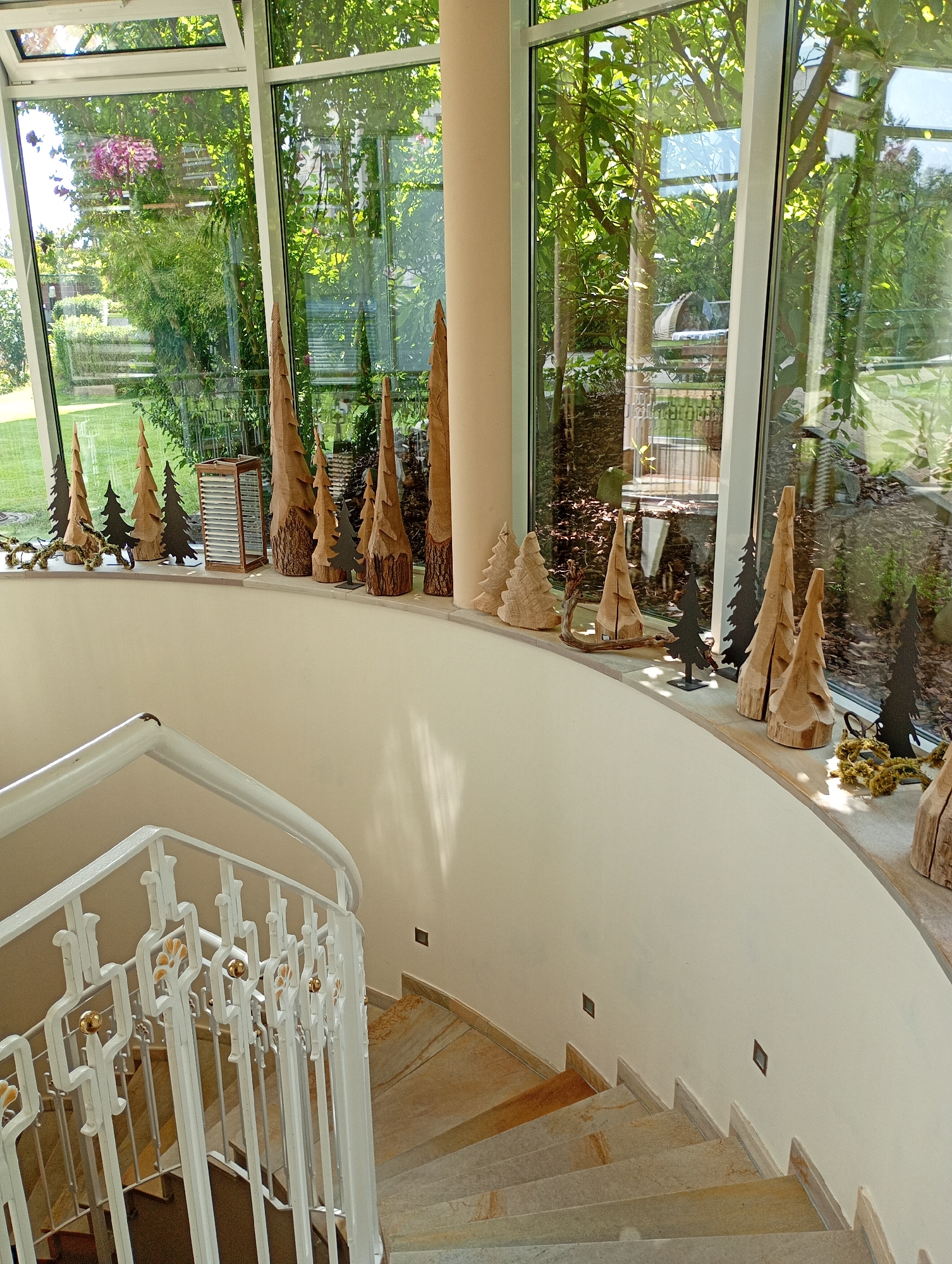 A high-angle, eye-level interior shot shows a curved staircase with a white railing and stone steps. The staircase is illuminated by natural light streaming through a large, floor-to-ceiling window that offers a view of a lush green garden. Along the windowsill above the staircase, a collection of wooden and metal decorative trees are displayed. The scene appears to be from a private residence or a hotel in Bad Sachsa, Germany.