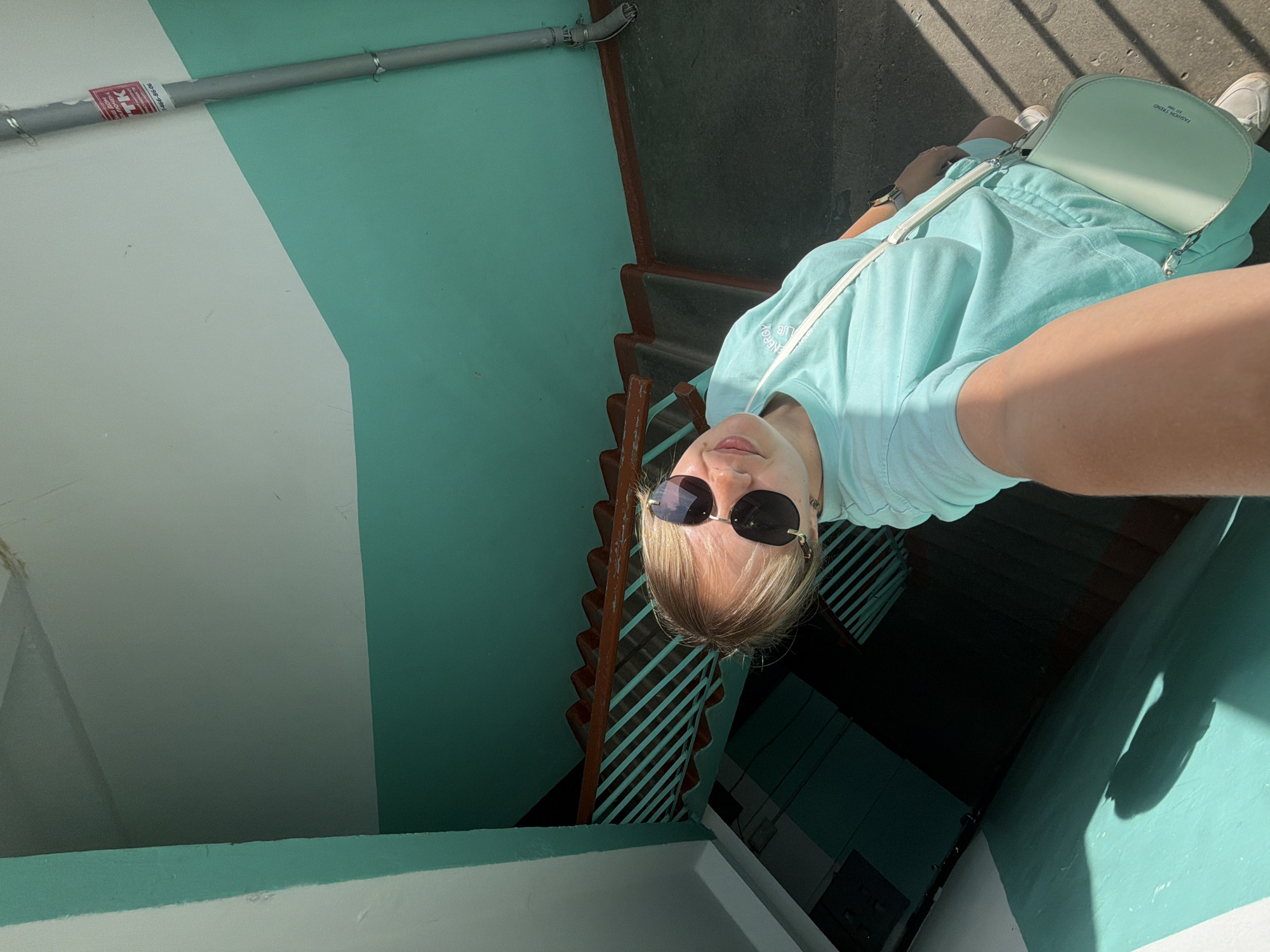 A woman takes a selfie on a staircase, holding the camera at arm's length. She wears sunglasses and a light-colored outfit. She is also carrying a light-colored handbag. The staircase is painted in two colors: teal and white. The building's interior shows elements of urban decay, with visible wear and tear. The lighting conditions suggest it is daytime. The image was taken in Chaikovsky, Russia.