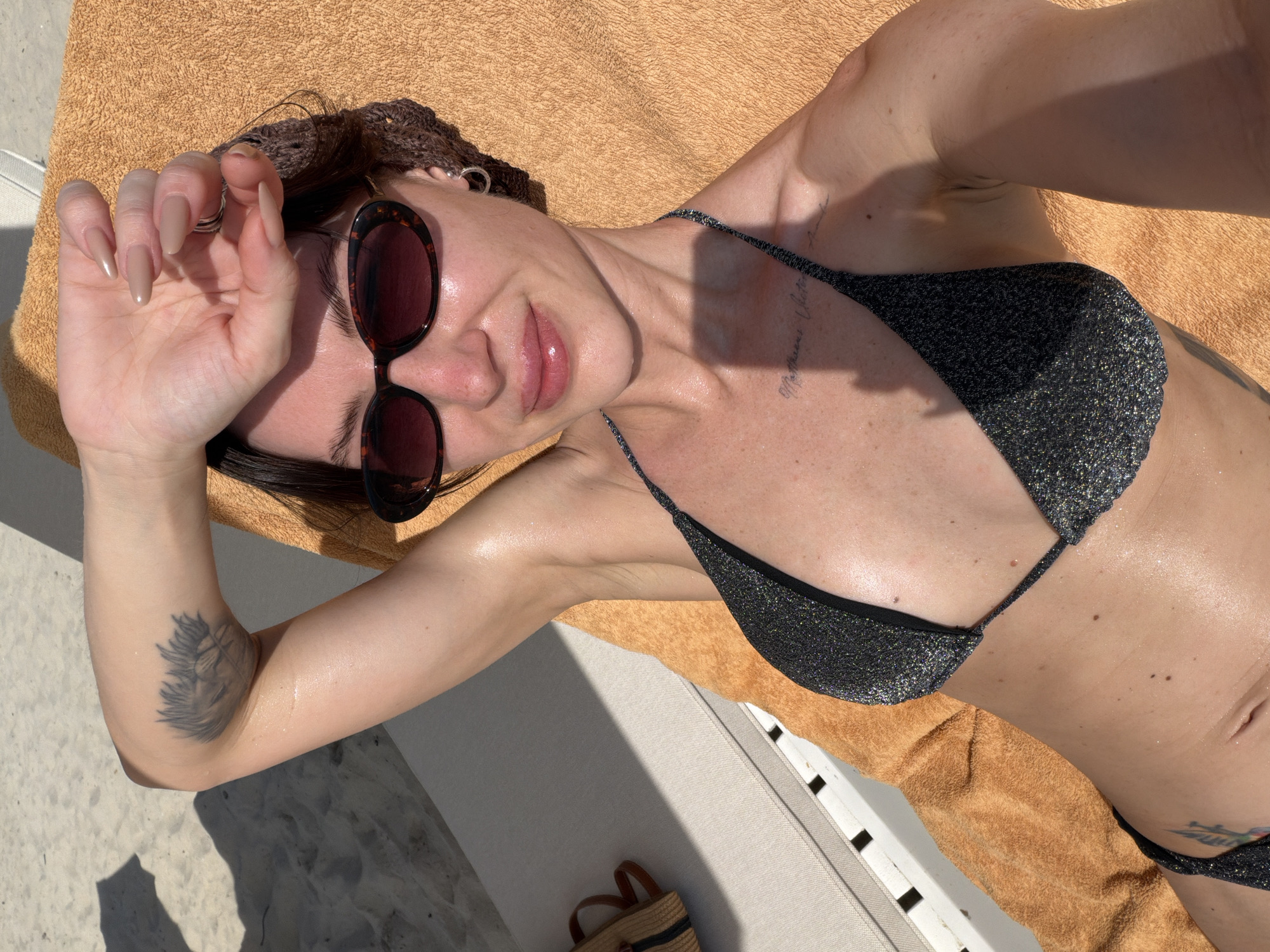 A female individual is observed sunbathing on a lounge chair in Dong Ninh Hoa Ward, Vietnam. She is positioned on an orange towel draped over a white slatted lounge chair. She has dark hair, partially covered by a brown crocheted head covering, and is wearing tortoise-shell framed sunglasses with dark lenses. Her right hand, featuring manicured, light brown, almond-shaped nails and a silver-toned ring on the middle finger, is raised to shield her face from direct sunlight. Her lips are glossy and she displays a slight smile. A small hoop earring is visible on her left ear.
The individual wears a dark, glittery bikini top. Visible tattoos include a stylized plant or pineapple design on the back of her left upper arm and script text on her upper left chest. Her skin appears glistening, consistent with the application of oil or sunscreen. In the background, light-colored sand is visible, indicating a beach or resort setting. Strong shadows from an unseen overhead structure are cast on the sand. A second white lounge chair is partially visible to the left, and a small woven bag with brown handles rests on the ground nearby.