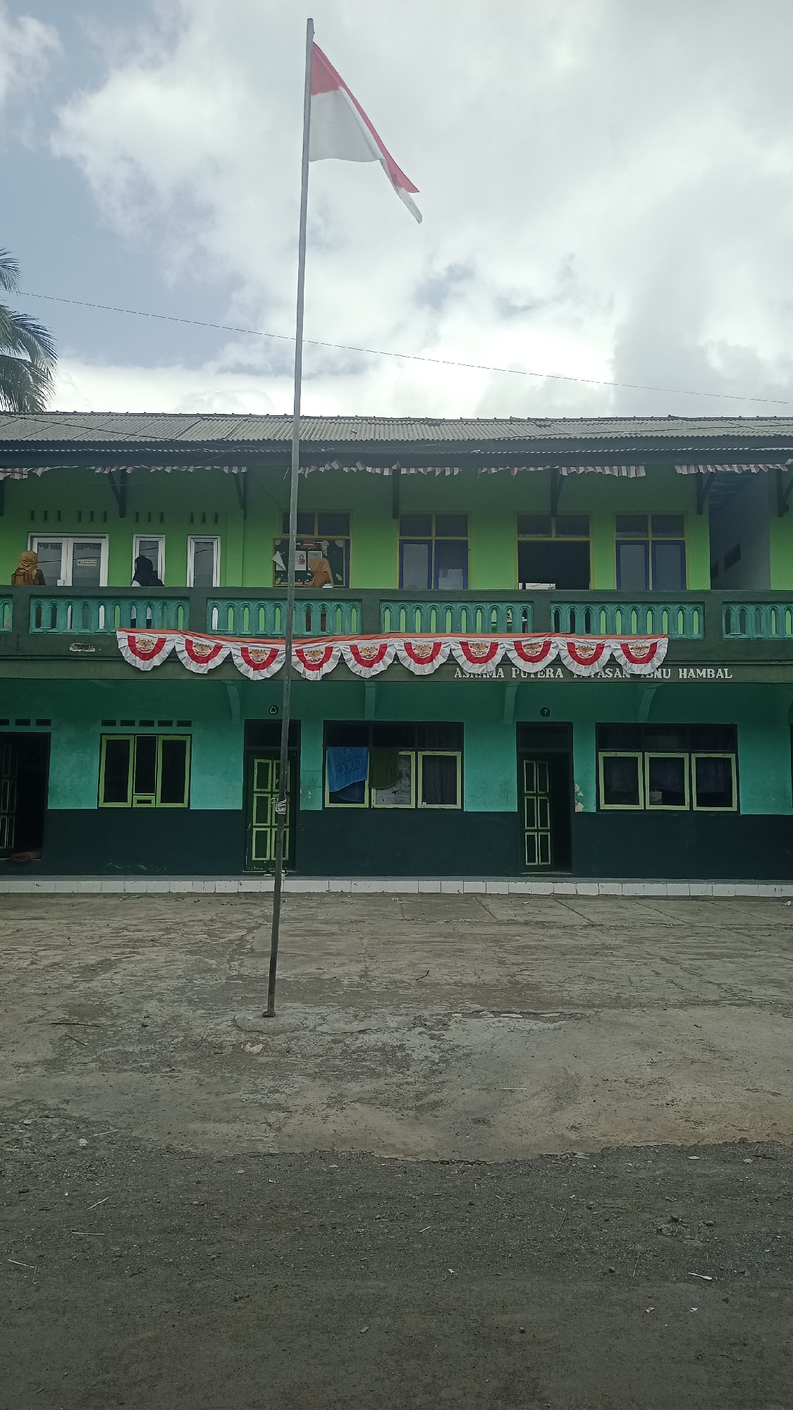 The image shows the exterior of a two-story green building, likely a school, located in Karyamandala, Indonesia. A flagpole with the Indonesian flag, red and white, stands prominently in front of the building. The building has multiple windows and doors, with some people visible on the second-floor balcony. A decorative banner with red and white triangular flags is hung across the second-floor balcony. The ground in front of the building is a paved courtyard area. The sky is cloudy, indicating it may be overcast or a partly cloudy day. The overall scene depicts a typical day at an educational institution in Indonesia.