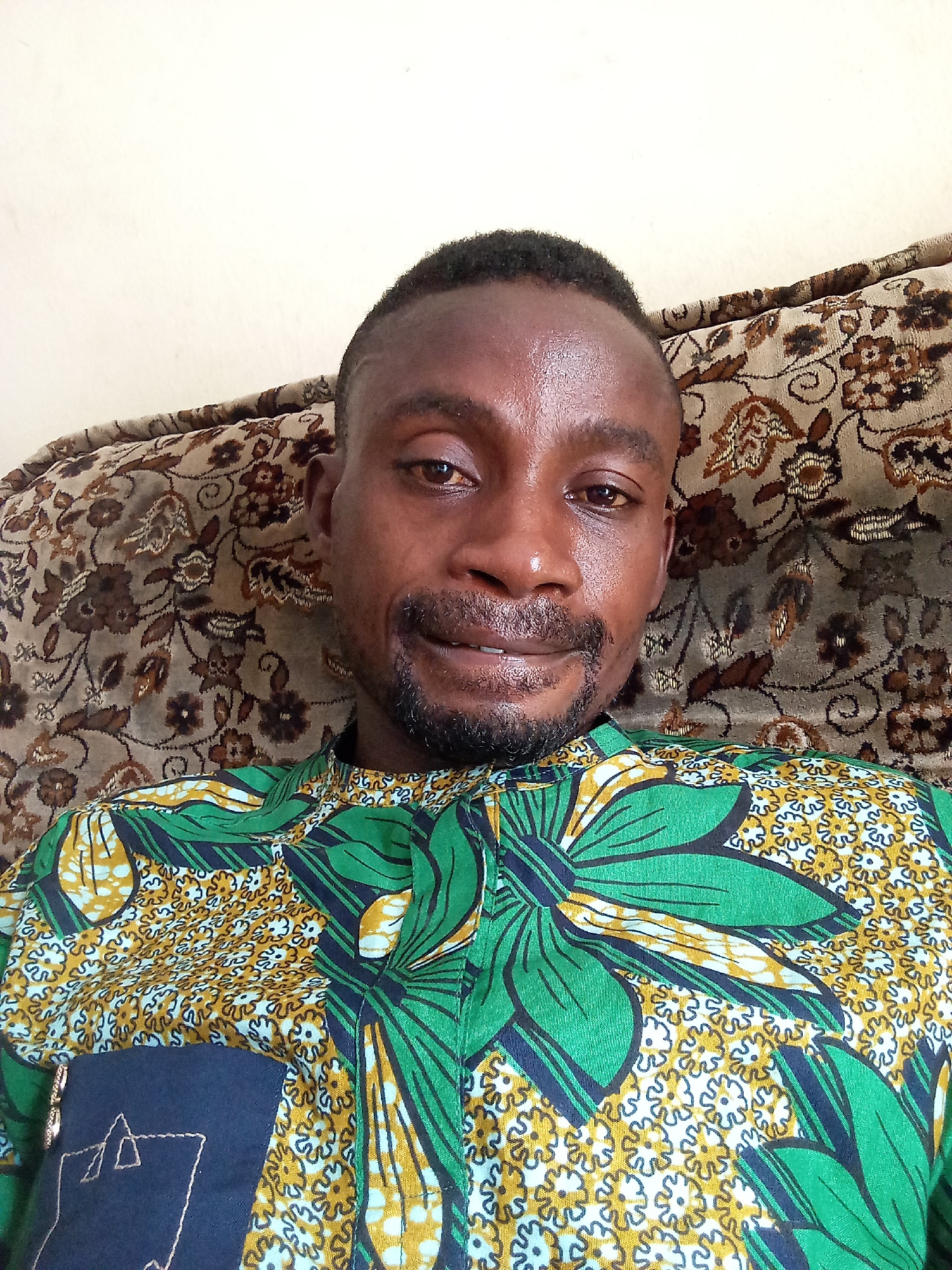 A man is shown from the shoulders up, positioned in front of a brown patterned sofa. He has dark skin, short dark hair, and a trimmed beard. His expression is neutral, and he seems to be looking directly at the camera. He is wearing a green and yellow patterned shirt with a navy blue patch pocket on one side. The photo appears to be taken indoors. There is no indication of a city.