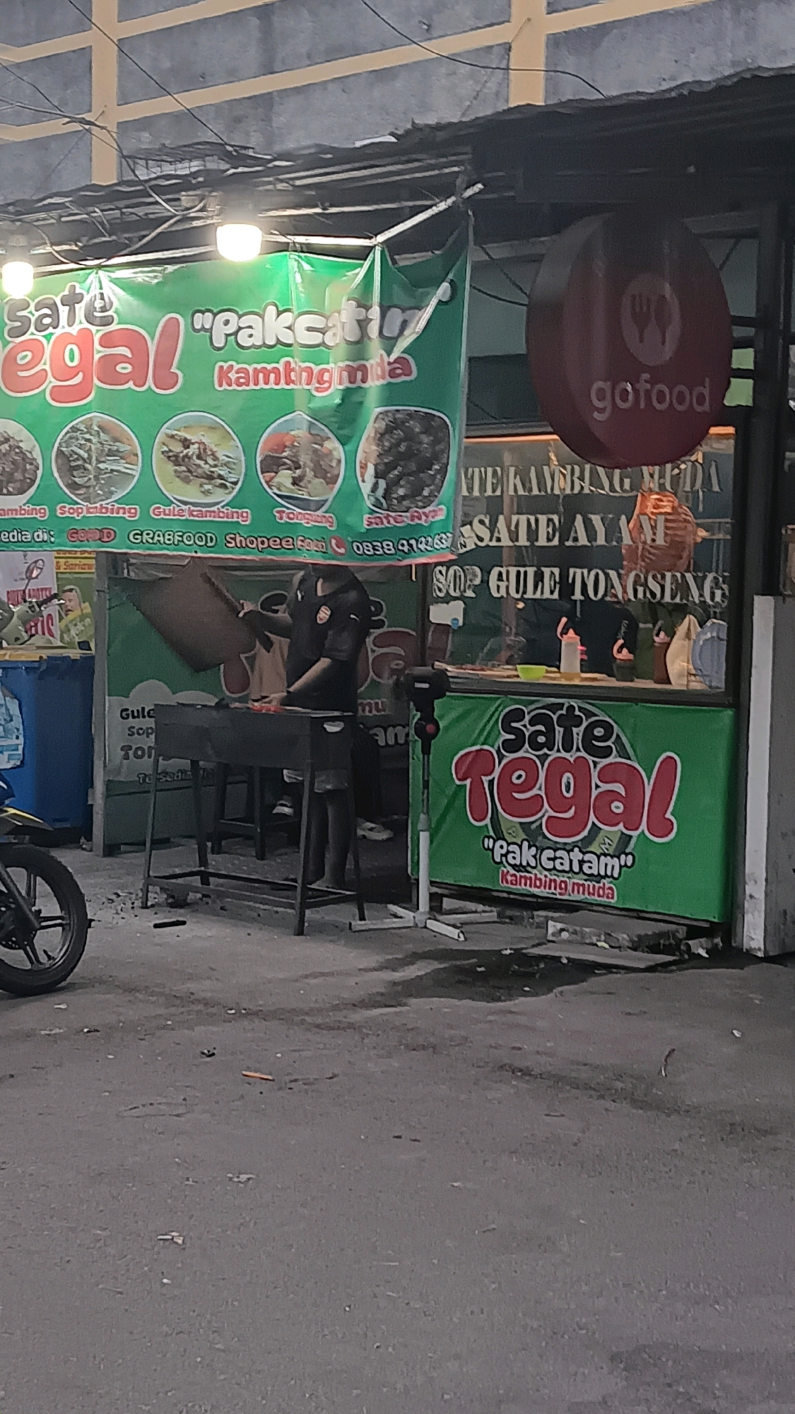 This is a medium shot, captured at ground level, of a street food stall in Depok, Indonesia. The stall, named "Sate Tegal", is adorned with vibrant green banners advertising various dishes. A man, wearing a dark uniform with a logo on the left breast, is standing behind a grill, holding a large utensil, and appears to be preparing food. The stall is equipped with a grill and is situated outdoors, likely on a street or sidewalk, given the asphalt ground. In the background, there's a glimpse of a building and a red GoFood sign, suggesting delivery services are available. The overall scene depicts a typical evening at a local Indonesian eatery.