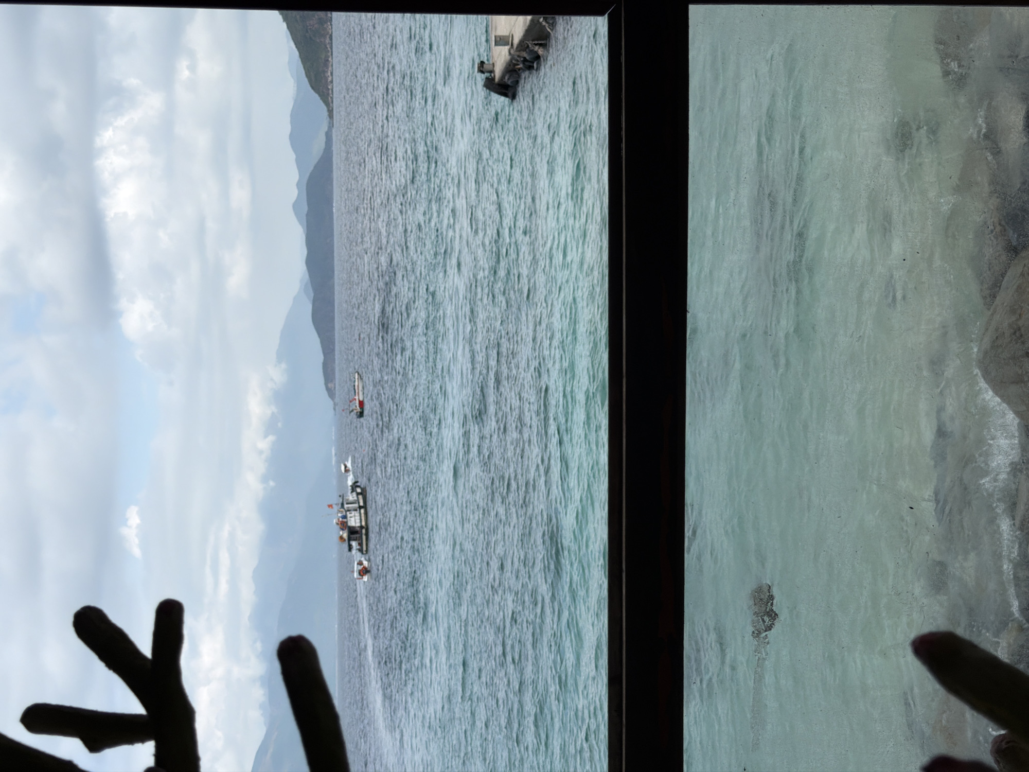 The image depicts a view from a window, likely in the city of Nha Trang Ward, Vietnam, looking out onto a body of water, possibly the sea. The water is choppy and appears a gray-blue color, suggesting cloudy or overcast weather. In the distance, there are mountains or hills visible on the horizon. Several boats are seen on the water, including one that appears to be a larger vessel, possibly a tour boat or fishing trawler, and a smaller boat closer to the frame. A dock is visible on top of the water. Silhouetted fingers are present in the bottom left and right corners, suggesting someone is holding or touching the window while taking the picture. The overall scene is of a coastal area with maritime activity and a somewhat overcast sky.