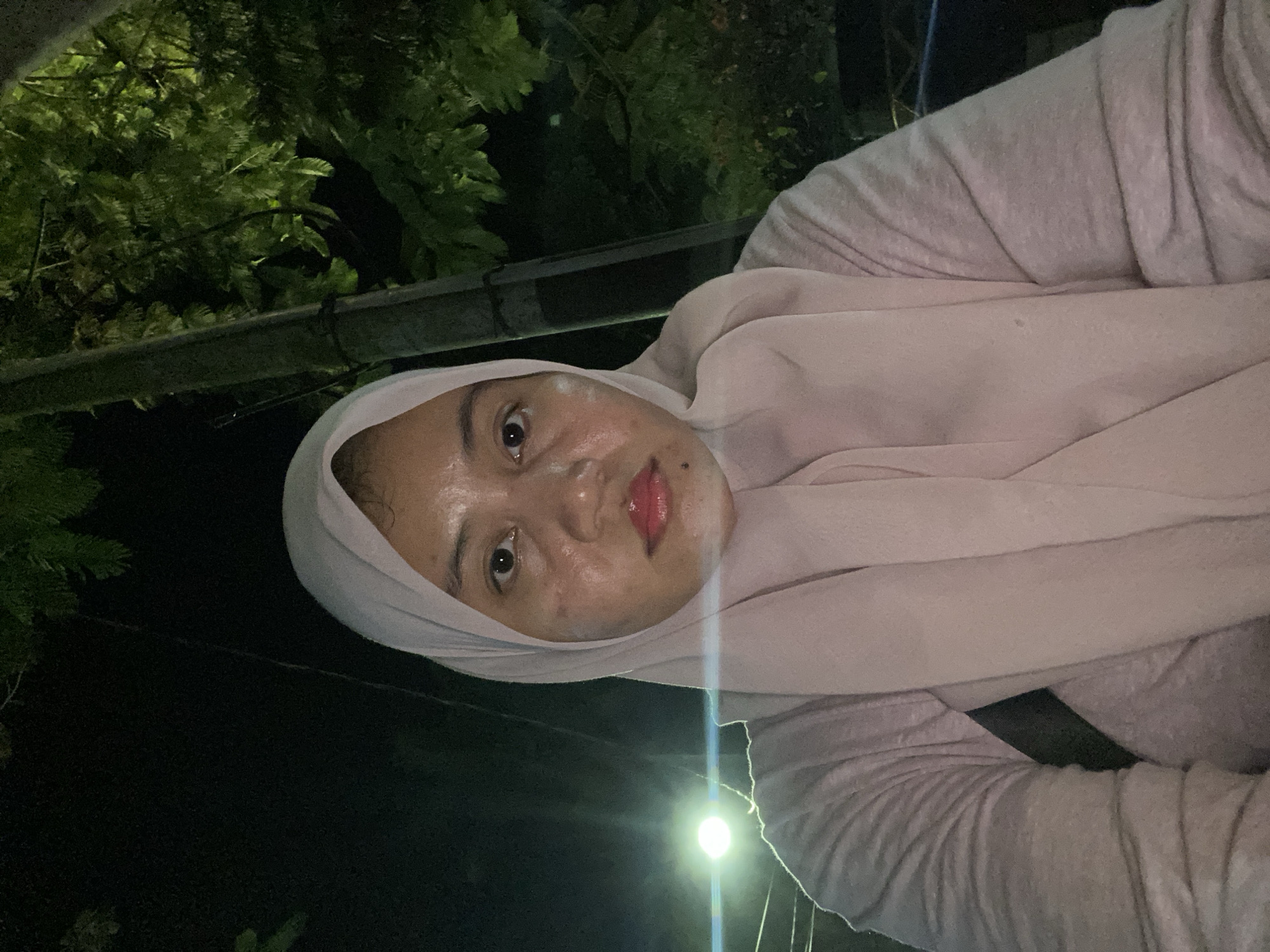 A young woman wearing a light pink hijab and a light pink top is photographed outdoors at night. She is looking directly at the camera. Her face is illuminated by a bright light source to her left. Behind her, a concrete pole is visible, and dense green foliage is to her right. The scene appears to be in a public outdoor space, possibly a street or park, in Banjarbaru, Indonesia.