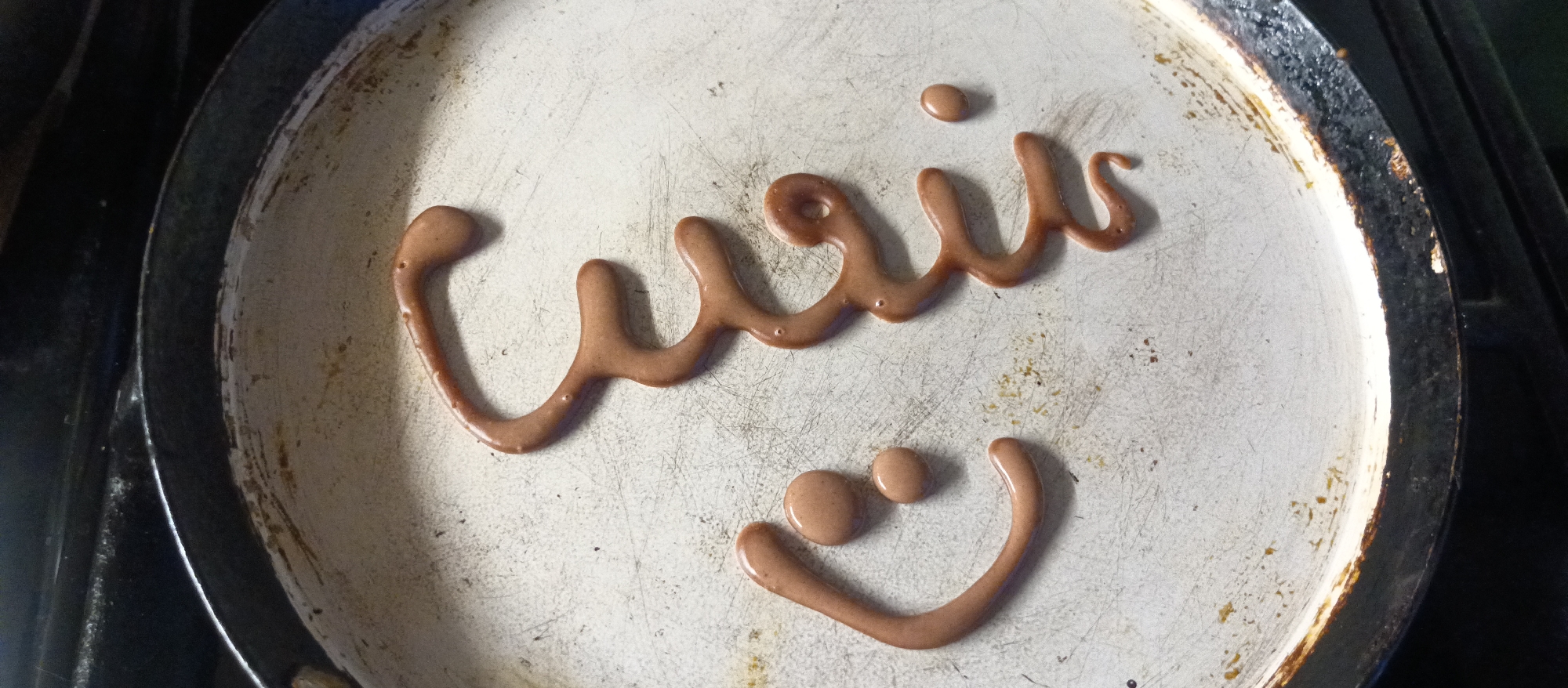 On a worn, off-white frying pan with a black rim, pancake batter has been used to write a word in Arabic script above a smiley face composed of two circular dots for eyes and a curved line for a mouth. The pan appears to be resting on a stove top, partially visible at the edges of the frame. The exact location is not discernible from the image.