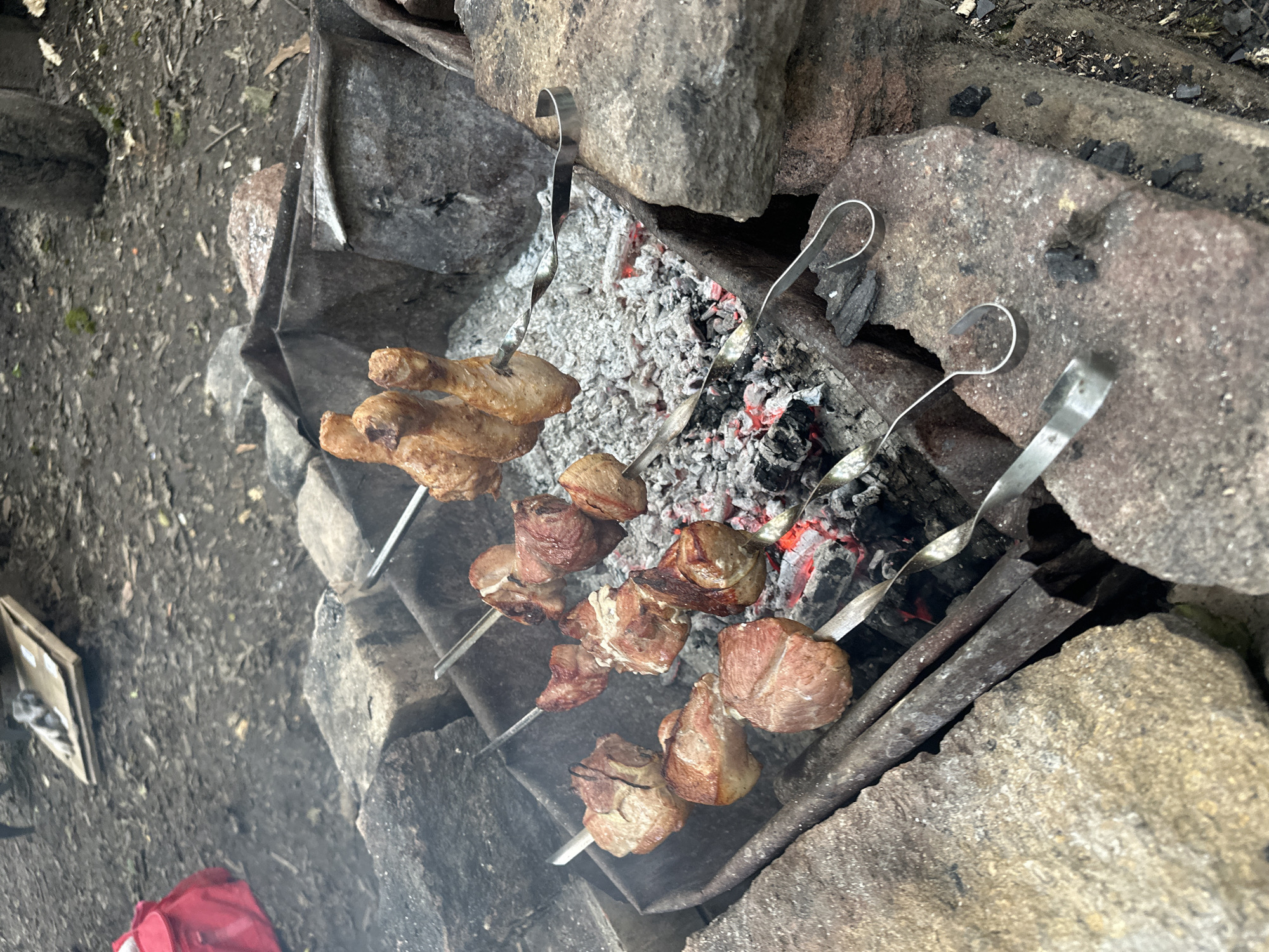 The image displays an outdoor cooking scene with multiple skewers positioned over glowing charcoal. Four metal skewers, two with twisted handles and two with simple hooks, hold food over a makeshift grill constructed from dark, possibly corroded, sheet metal sections contained by various rough-hewn stones. Three of the skewers on the left hold multiple irregularly shaped chunks of light brown, seared meat, consistent with pork kebabs (shashlik). The skewer on the far right holds three browned chicken drumsticks. Red-hot coals and white ash are visible beneath the food, with faint smoke rising. The ground surrounding the grill is dark, uneven soil with scattered debris. In the upper left background, a red bag is visible on the ground, next to a flattened cardboard box topped with a grey textile item. A dark, cylindrical object, possibly a tree stump, is partially visible in the upper right background. No individuals are present in the scene. The location is an outdoor, rustic environment in Unknown, Unknown.