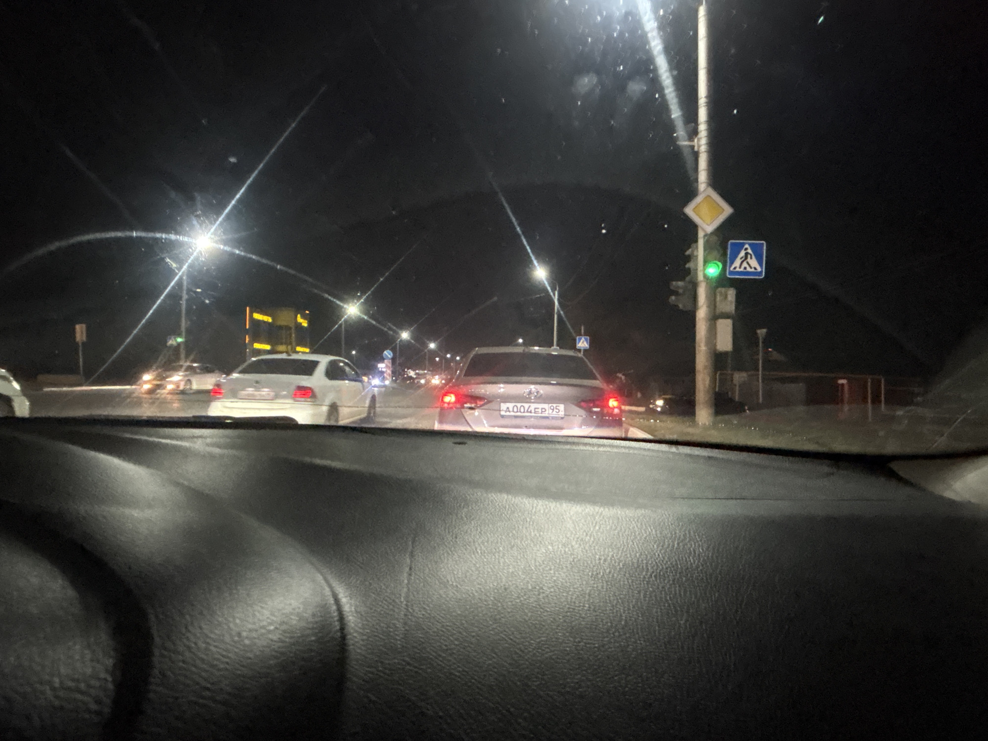 The image captures a nighttime scene from the interior of a vehicle, looking forward through the windshield. The view includes a traffic intersection at night in the city of Grozny, Russia. In the foreground, the textured black dashboard of the car is visible. Beyond the dashboard, the road and surrounding environment are illuminated by streetlights and vehicle lights. Several cars are present on the road, with their headlights and taillights visible. One car in the middle ground, a light-colored sedan, has its rear taillights illuminated and a license plate that reads "A004EP 95". To the right of this car, a traffic light shows a green signal, and adjacent to it are traffic signs: a yellow diamond-shaped sign and a blue square sign depicting a pedestrian crossing. Further down the road, more vehicles and streetlights are visible, creating a typical urban night traffic scene.