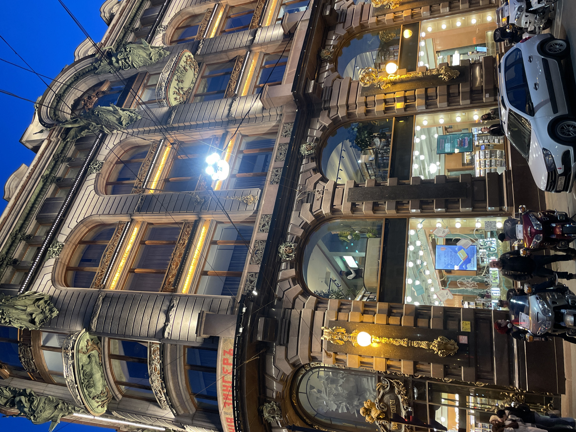 An upward-angled view captures the ornate facade of a multi-story building at dusk or night in Saint Petersburg, Russia. The architecture combines elements of classical and Art Nouveau styles, featuring numerous arched windows, decorative cornices, and sculptural details. Prominently displayed on the upper levels are dark, green-patinated dragon or griffin-like figures, adding to the building's distinctive character.
The sky is a deep twilight blue, contrasting with the warm, artificial lighting illuminating the building. On the lower levels, large display windows showcase brightly lit interiors, typical of commercial establishments. Decorative golden streetlights with ornate bases are affixed to the facade, casting a warm glow. Reflections of people and indoor lights are visible in the windows.
On the street level, a white car is parked in front of the building, alongside several motorcycles. People are visible on the sidewalk, appearing to be walking or looking into the storefronts. Overhead power lines crisscross the upper part of the image.
Visible text on a blue sign in a storefront window reads "ВСЁ ЛЕТО СКИДКИ" (ALL SUMMER DISCOUNTS), indicating a retail business. Another partially visible sign below a golden decoration on the lower facade appears to show "РИНГ" (RING). The scene depicts a lively urban evening atmosphere.
