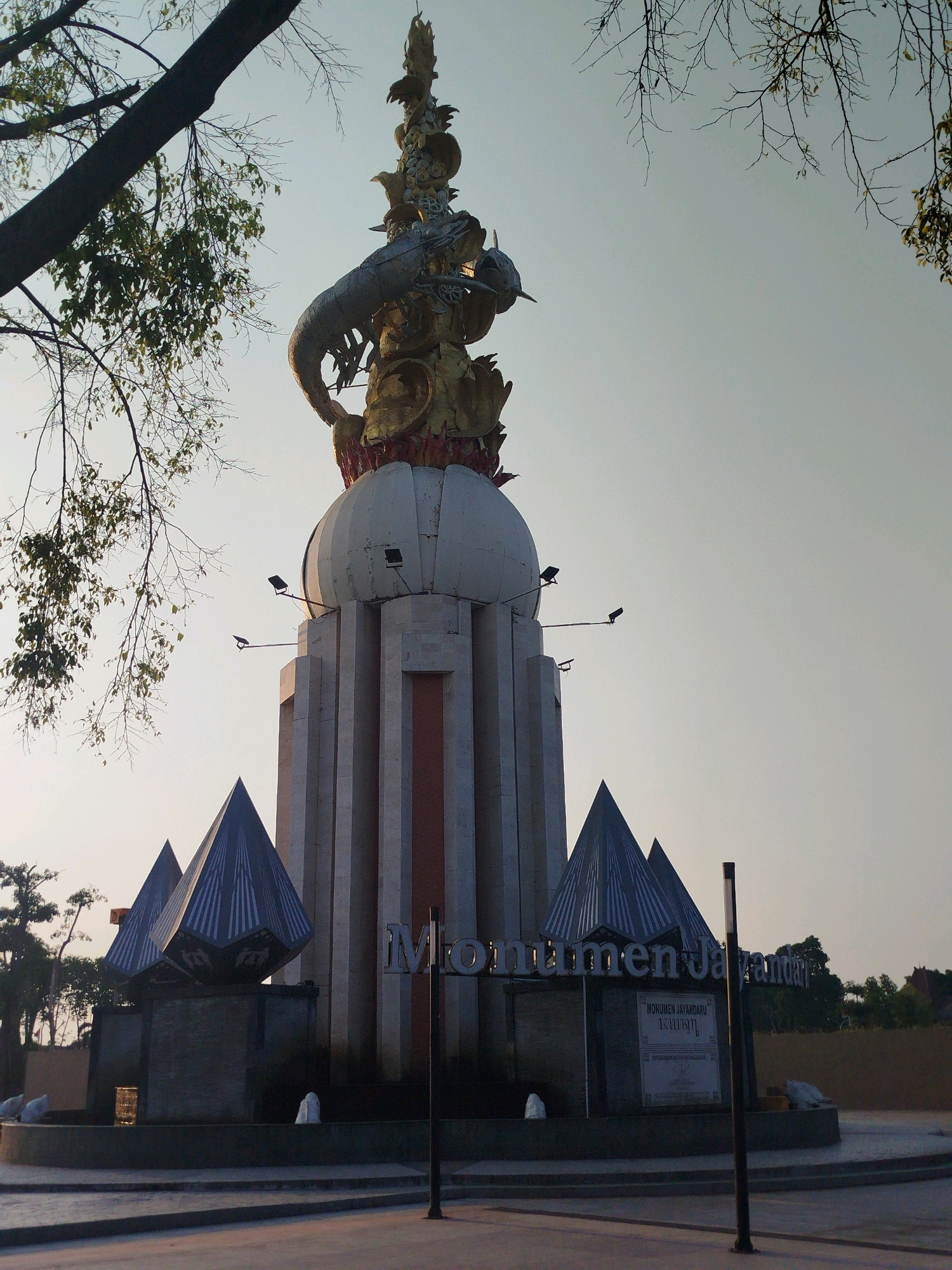 This is a low-angle, full shot of the Jayandaru Monument in Sidoarjo, Indonesia, bathed in the soft light of either dawn or dusk. The monument is the central subject, a towering structure with a striking, ornate golden sculpture at its apex, featuring a large serpent-like creature wrapped around a smaller dragon-like figure. Below this, a white, spherical section connects to a tall, rectangular base composed of pale, vertical stone pillars, accented by reddish-orange vertical panels. Flanking the base are two large, dark, pyramid-shaped structures, their surfaces adorned with intricate white geometric patterns. In front of these pyramid structures, a fountain area is visible, with white stone figures and water jets. The monument bears the inscription "Monumen Jayandaru" in large, metallic letters. To the right, a plaque with Indonesian text is mounted on a dark signpost. In the foreground, a paved walkway leads towards the monument, and a street is visible. Trees with bare branches and some sparse green leaves frame the top of the monument and the sky. The sky itself is a pale, clear blue, indicating a calm atmosphere. There are no people visible in the scene.