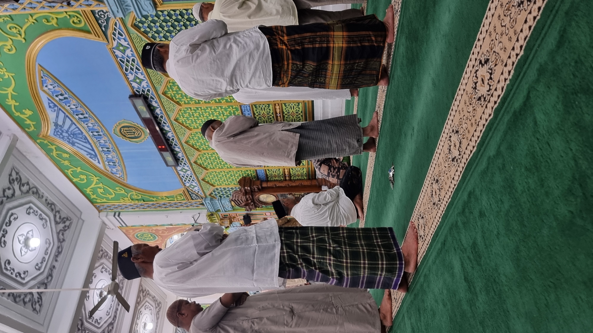 The image depicts a group of men engaged in prayer inside a mosque in Keudekruenggeukueh, Indonesia. The men are dressed in traditional Islamic attire, including sarongs and caps. They are standing and kneeling on a green carpeted floor, facing a wall adorned with intricate green and blue decorative patterns. A large wooden clock is visible on the left side of the wall. The ceiling features ornate white decorative plasterwork and a ceiling fan. The overall atmosphere suggests a religious gathering.