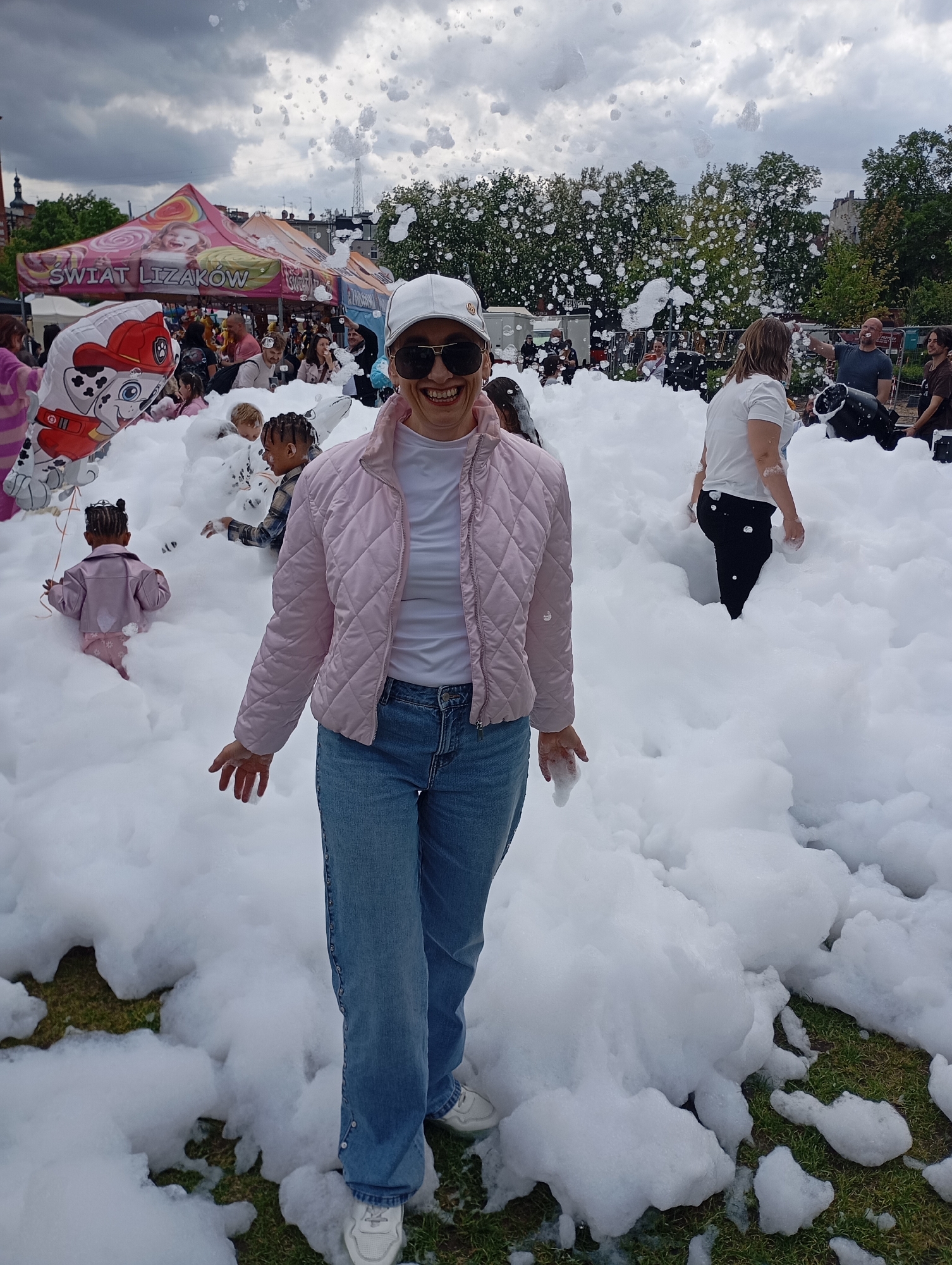 A woman stands smiling in a sea of foam at a public event likely in Bydgoszcz, Poland. She wears a white baseball cap, sunglasses, a pink quilted jacket, a white t-shirt, blue jeans, and white sneakers. The foam reaches up to her knees. Behind her are other people, including children, playing in the foam. To the left is a "Świat Lizaków" tent. In the background are trees and buildings, typical of an urban environment. A balloon with Marshall from Paw Patrol is visible among the people. A man operating a foam cannon can be seen to the right.