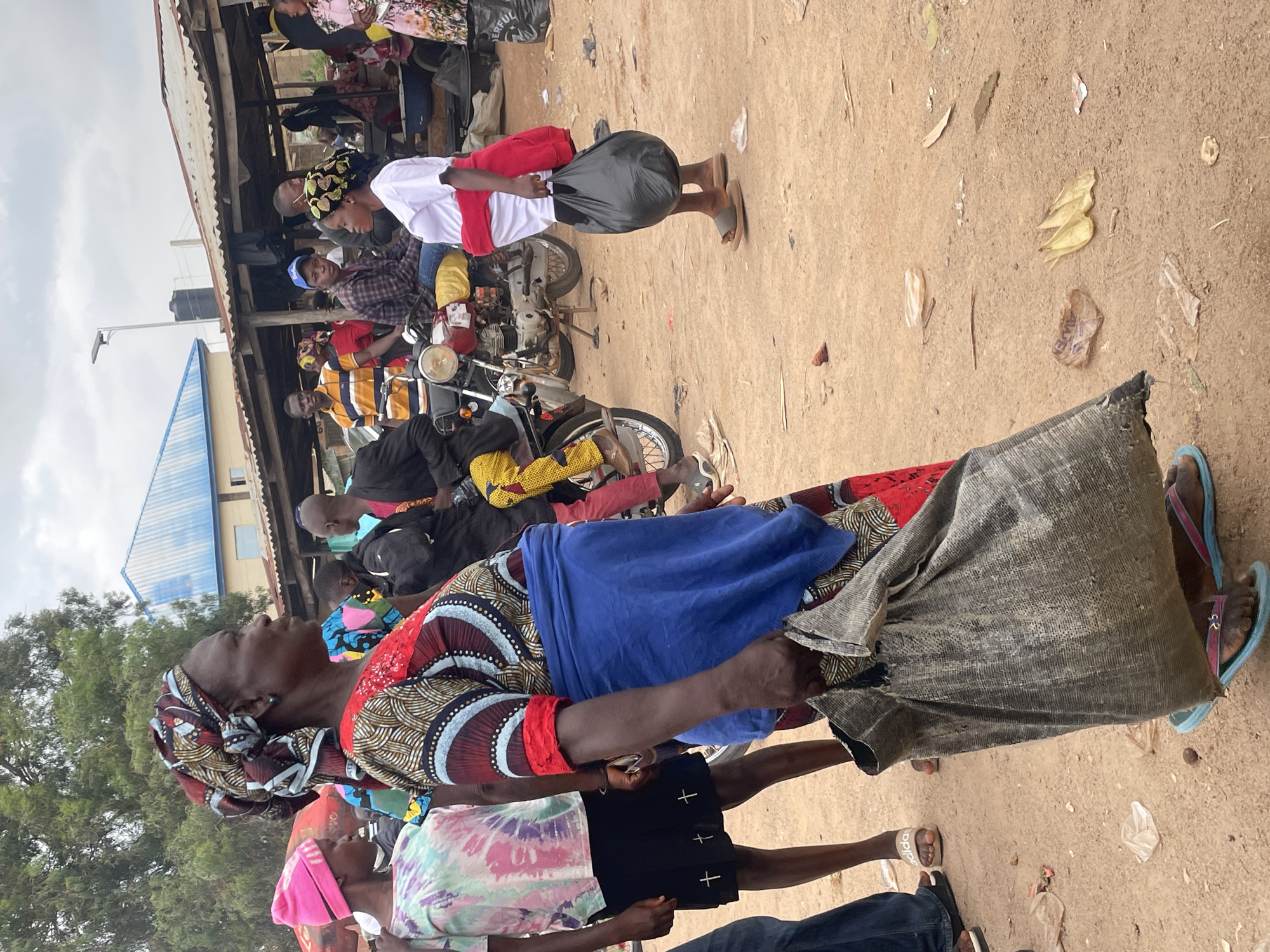 The image depicts a bustling outdoor market scene in Wang, Nigeria. In the foreground, a woman wearing a colorful patterned dress and a headscarf carries a large, rough woven sack. To her left, another woman in a tie-dye t-shirt and black skirt is visible. Behind them, several individuals are gathered around motorcycles and market stalls. One stall has a corrugated metal roof, and various goods are displayed. The ground is made of packed dirt, with scattered debris and some dried plant matter. The atmosphere appears lively, with people engaging in commerce and movement. The sky is overcast.