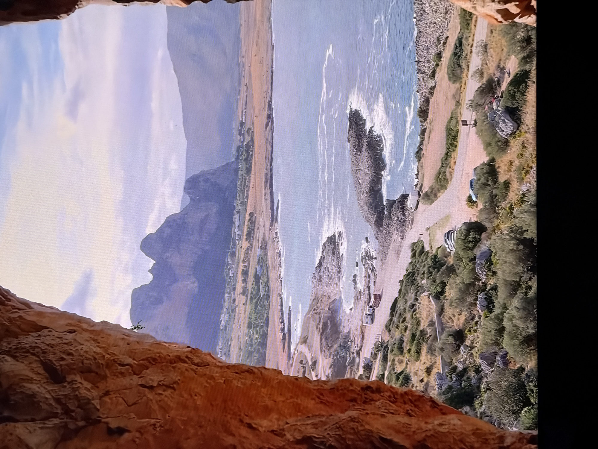 The image displays a scenic coastal landscape viewed from within a rocky cave opening. In the foreground, the rough, textured orange-red rock of the cave entrance frames the scene. Beyond the cave, a body of water, likely the sea, stretches out, with gentle waves breaking against a rocky shoreline. A winding road traverses the coastal terrain, dotted with sparse vegetation and some vehicles, including what appear to be campers or recreational vehicles. In the distance, rolling hills and mountains rise under a sky filled with soft clouds. The overall impression is one of a natural, tranquil setting. The image does not depict the city of Chirchik, Uzbekistan.