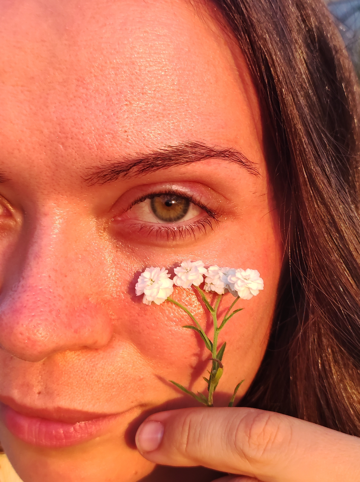 The image is a close-up portrait of a person's face, with a focus on their eye and cheek. A delicate stem with small white flowers is held near the cheek, with a finger subtly visible in the foreground. The lighting suggests it is either sunrise or sunset, casting a warm, orange glow across the skin and hair. The background is out of focus, with hints of natural elements like foliage or sky. The setting is likely outdoors, as indicated by the natural light and the presence of the flower. No specific location in the city of Фоки, Russia can be determined from the image itself.