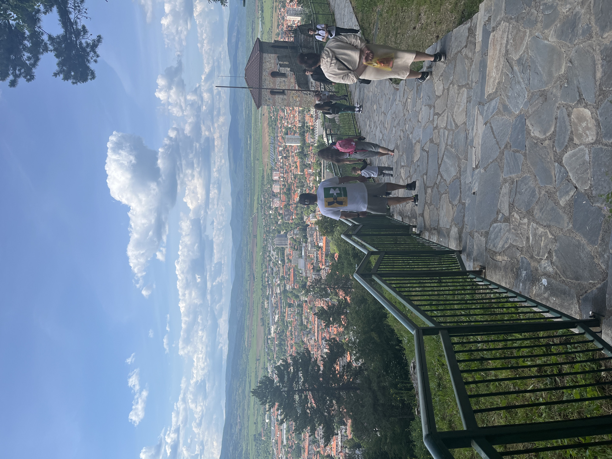 The image captures a scenic overview of Kyustendil, Bulgaria, from what appears to be a high vantage point. The viewer looks down over a city with densely packed orange-roofed buildings stretching out towards a green valley and mountains in the distance under a blue sky populated with puffy white clouds. In the foreground, there's a stone pathway with a metal railing. Several people are present, including a man in a white T-shirt with a logo on the back, walking downhill on the path. Other people are observed near a small brick building with a brown tiled roof. The location is likely a popular viewpoint or tourist spot offering panoramic city views.