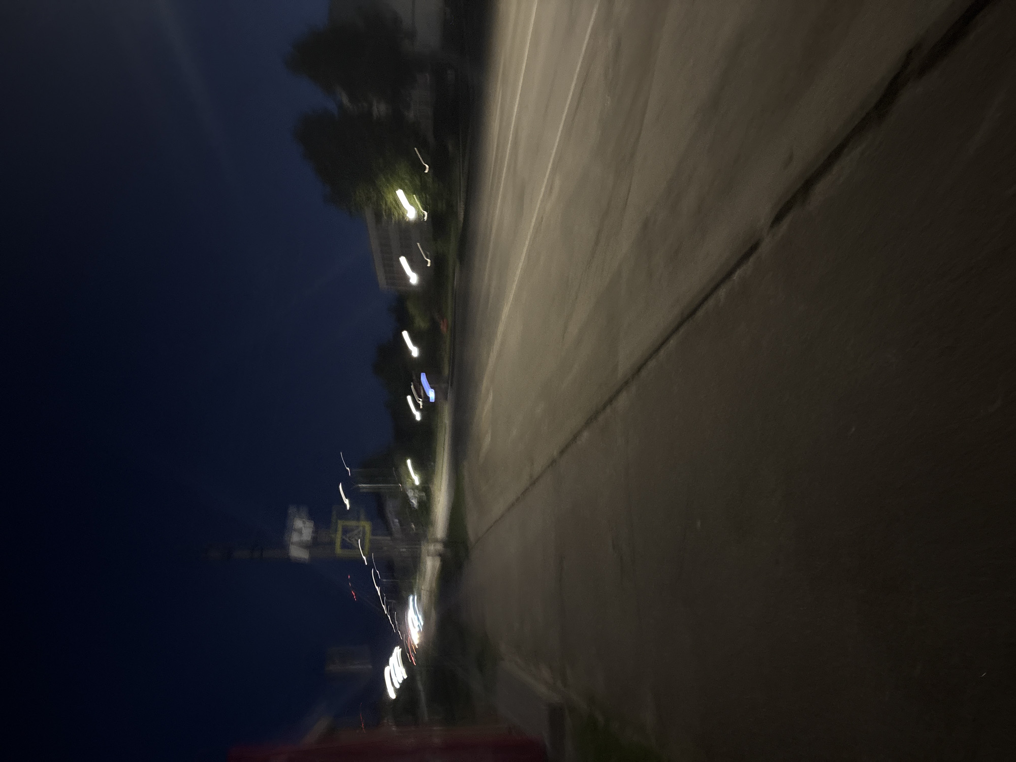 The image depicts a nighttime street scene in an unknown urban or suburban location. The scene is characterized by significant motion blur, suggesting either a moving camera or a long exposure photograph. In the foreground on the left, a dark paved sidewalk is visible, running parallel to a multi-lane paved road. The road features indistinct white lane markings. Along the street, luminous white and red light trails, indicative of moving vehicle headlights and taillights, streak horizontally across the midground. A yellow diamond-shaped pedestrian crossing sign is mounted on a pole on the left side of the road. Additional poles, likely for streetlights or utilities, are scattered throughout the image. Silhouetted trees and blurry building outlines are discernible in the background. The sky is a dark navy blue, consistent with nighttime conditions. No individuals are present in the frame. A reddish structure is partially visible on the far left edge of the image.