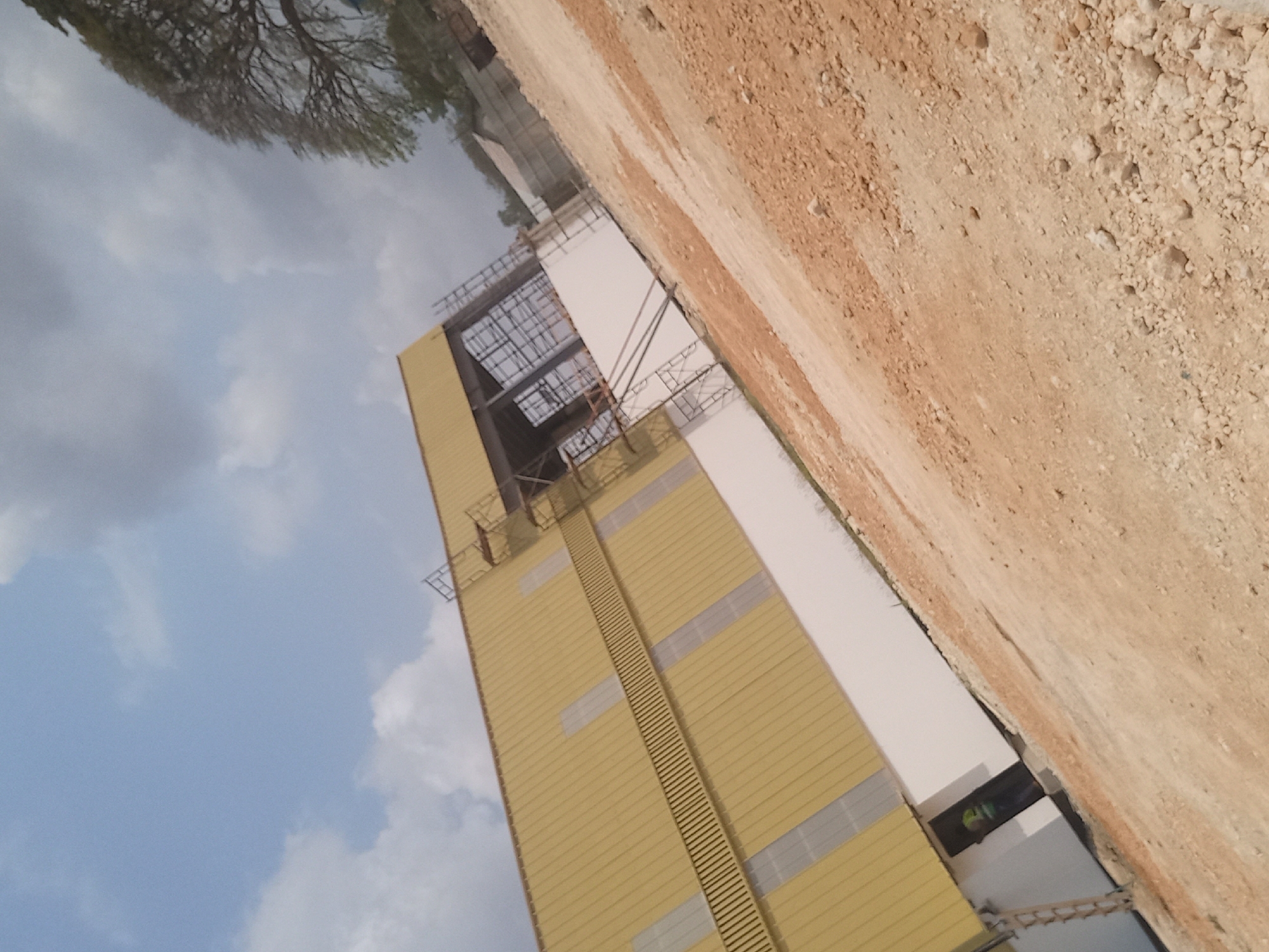The image depicts a construction site in Bububu, Tanzania. A large, yellow, metal-clad building is partially constructed, with scaffolding visible on its side. The building has several rectangular windows that appear to be tinted or covered. A white base runs along the lower portion of the building. The surrounding area is a dirt construction site with loose soil and small rocks. A person wearing a high-visibility vest is visible near a dark doorway on the side of the building. In the background, a large tree with lush green foliage is present, along with other structures that suggest an ongoing development. The sky is partly cloudy with a mix of white and grey clouds. The image is taken from a slightly elevated, tilted angle, looking upwards and towards the building.