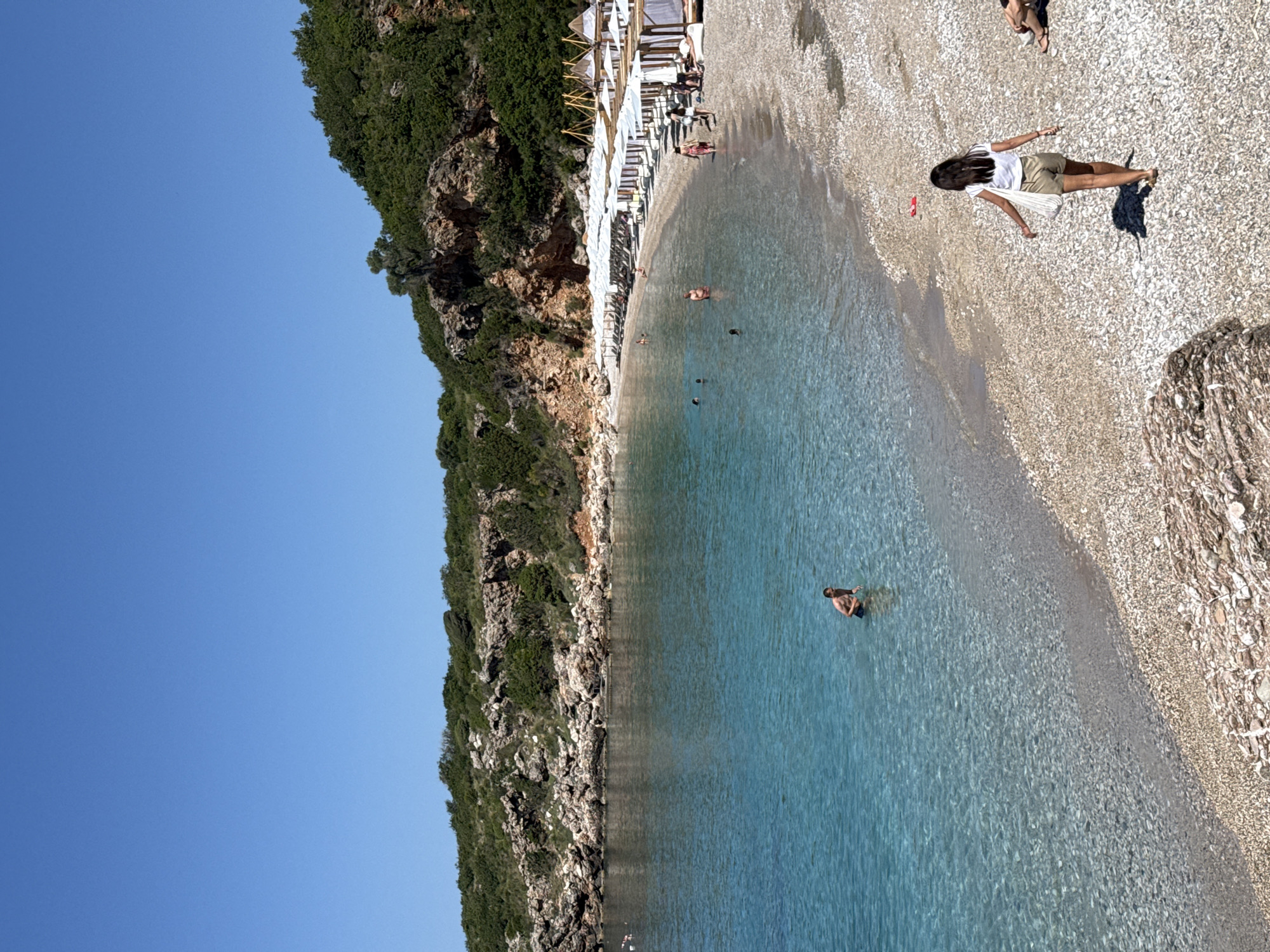 The image depicts a pebbled beach cove in Drobnići, Montenegro, under a clear, bright blue sky. The cove is flanked by rocky hills covered in green vegetation. The water in the cove is transparent and turquoise, revealing the pebbly seabed near the shore.
On the right side of the beach, numerous white beach umbrellas are arranged in rows, each accompanied by lounge chairs. Further to the right, several larger cabanas with white fabric canopies and wooden frames are visible.
Several individuals are present. In the foreground, a woman with dark hair, wearing a white t-shirt and light-colored shorts, walks away from the viewer on the pebbled beach, carrying a light-colored tote bag over her shoulder. A small red object rests on the pebbles near her. In the water, a shirtless male stands chest-deep towards the left of the frame. Other individuals are partially submerged or swimming further out in the bay. On the beach, people are visible sitting on lounge chairs under umbrellas and walking near the water's edge and cabanas.