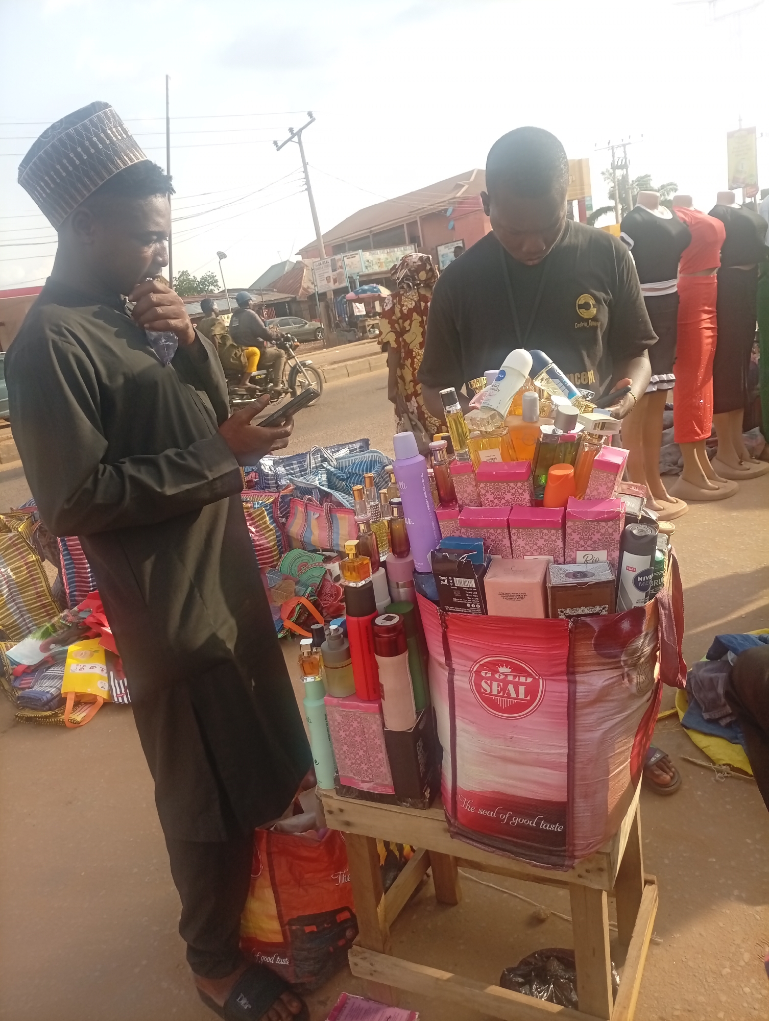 In an outdoor scene in Sabon Tasha, Nigeria, two men are standing near a collection of merchandise displayed on a small wooden table and on the ground. The man on the left, wearing a dark robe and a patterned hat, is looking at a mobile phone in his hand. The man on the right, dressed in a dark t-shirt with a logo, is looking down at the displayed items. The merchandise consists primarily of perfumes, deodorants, and lotions, neatly arranged in boxes and bottles, packed in a "GOLD SEAL" bag. Mannequins wearing dresses and shirts are in the background. There is a street with a motorcycle and buildings visible in the distance. Bags, possibly containing other goods, are scattered to the left of the table.