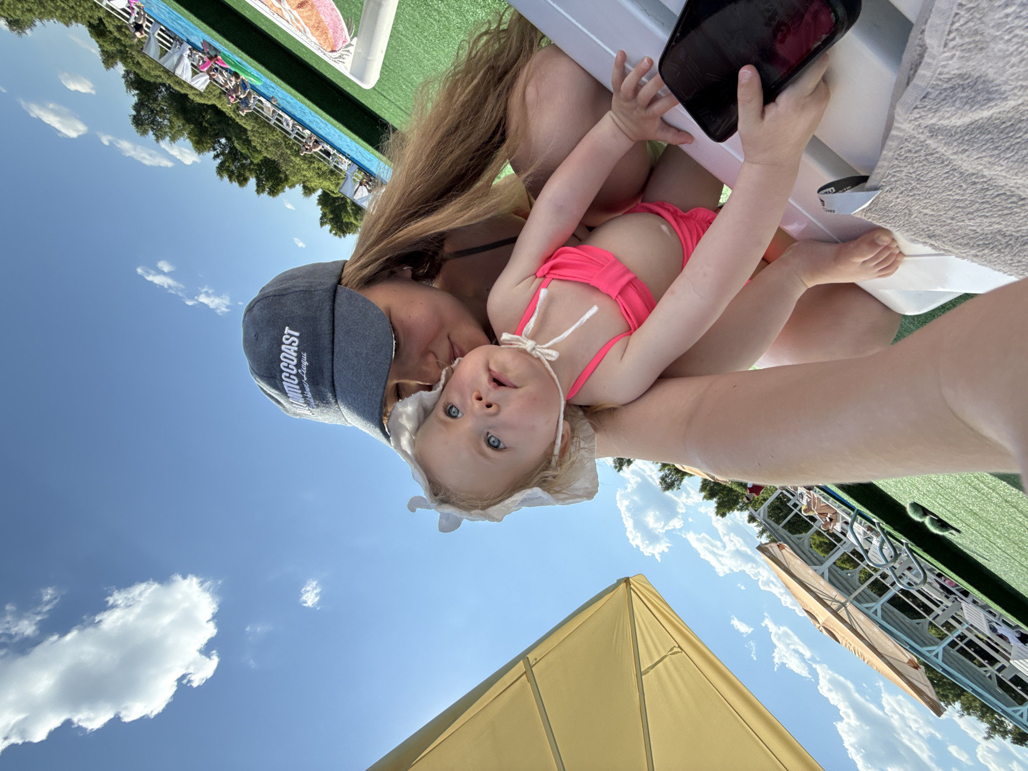 A woman wearing a gray baseball cap and a pink bikini holds a baby wearing a white bonnet and a pink bikini. They are sitting on a white lounge chair outdoors. The background shows a blue sky with white clouds, green grass, trees, and a swimming pool with people in it. A yellow umbrella is also visible. A phone is held in the woman's right hand, partially covering the baby's leg. The scene appears to be at a water park or resort in Koloksha, Russia.