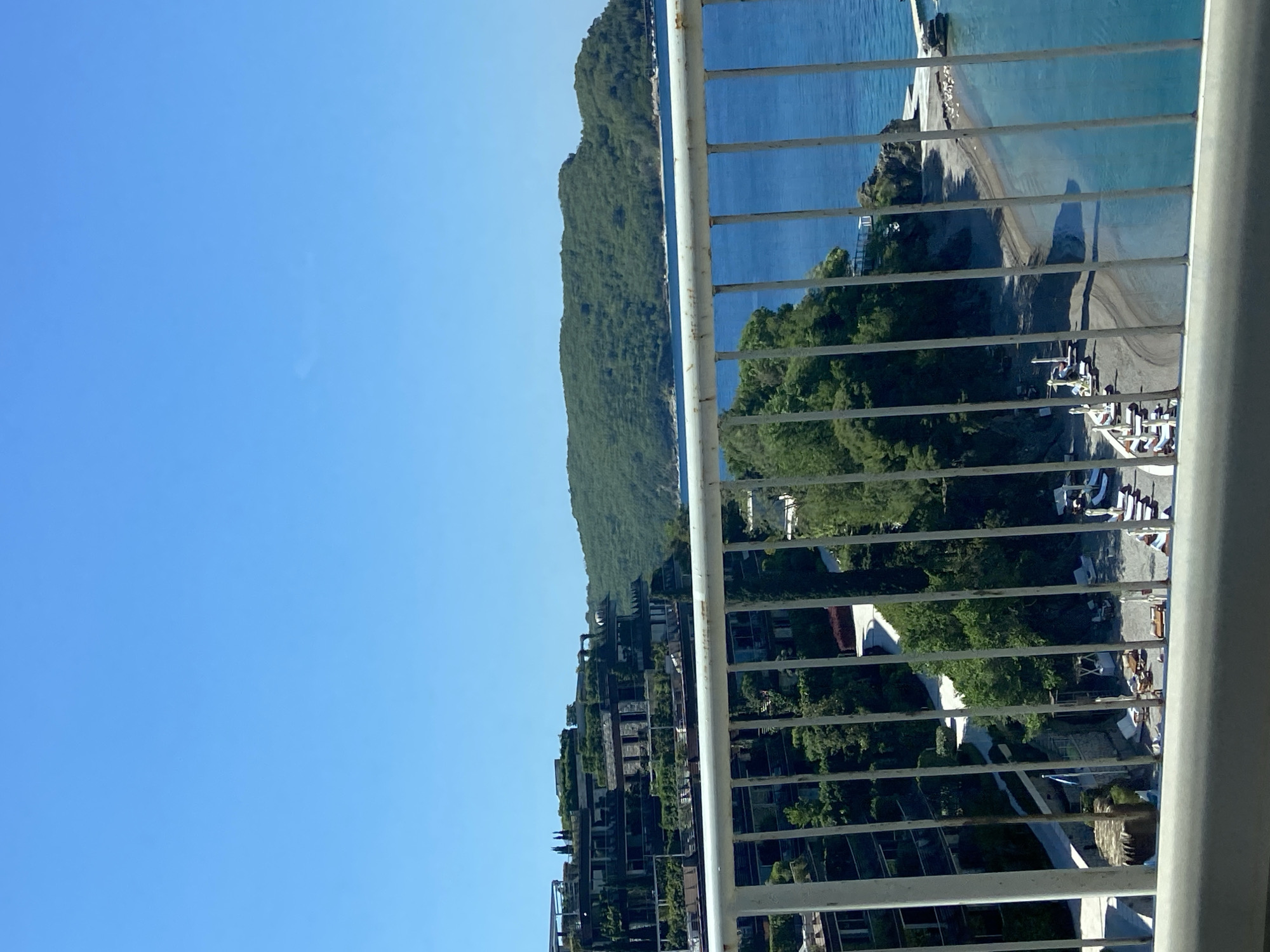 This panoramic image captures a vibrant coastal scene, likely in Budva, Montenegro, viewed from a high vantage point, such as a balcony. In the immediate foreground, a white metal balcony railing, consisting of vertical bars and a horizontal top rail, frames the view.
The scene unfolds with a clear, azure sky dominating the upper portion of the frame. Below the sky, lush, green, tree-covered mountains or hills extend along the coastline. To the right, a calm, turquoise sea stretches out, meeting a sandy or pebbly beach. On the beach, a long, distinct shadow, possibly cast by a person or a structure, stretches across the sand. Scattered on the beach and an adjacent paved area are various white objects, possibly sun loungers, umbrellas, or beach resort furniture, indicating a recreational area.
Further inland, nestled into the lower slopes of the hills, are several modern buildings, resembling a resort or hotel complex. These structures are multi-storied, with balconies and terraces, and are interspersed with green foliage, suggesting integrated landscaping or rooftop gardens.
The time of day appears to be sunny, with strong, bright light and discernible shadows, indicating mid-morning or late afternoon. The weather is clear and cloudless. There is no discernible specific activity taking place, though the presence of resort amenities implies leisure. No visible text is present in the image.