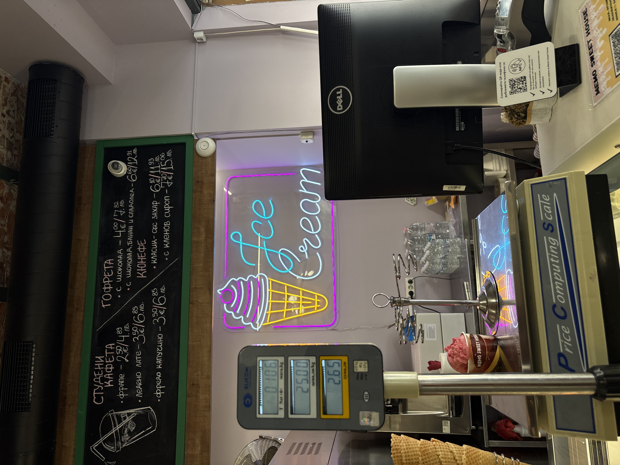The image shows the counter of an ice cream shop, likely in Plovdiv, Bulgaria. A black chalkboard on the left lists the menu items and prices in both local currency and euro. A neon sign reads "Ice Cream" above a scale displaying a weight of 0.106 kg, a price per kg of 25.00, and a total cost of 2.65. There is an ice cream cone on the right side of the scale. A Dell monitor is mounted above the counter. The words "Price Computing Scale" are also visible in the shot.