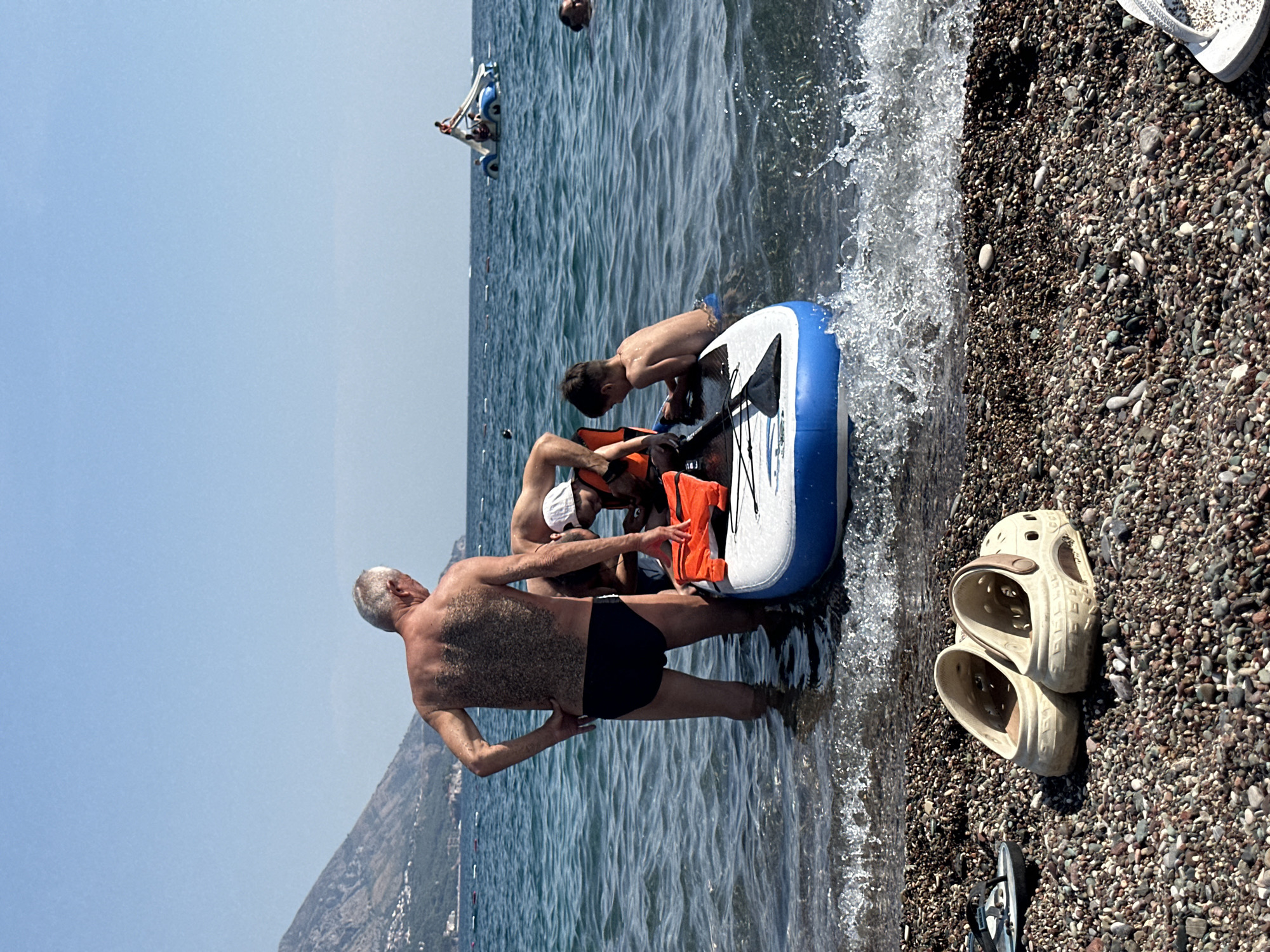 In the city of Boreti, Montenegro, on a sunny day, an elderly man with gray hair and a hairy back stands in shallow, clear blue water near a pebbled beach. He is wearing black swimming trunks. To his right, several people are gathered around an inflatable paddleboard. An adult, partially visible, is assisting a young boy wearing an orange life vest who is kneeling on the paddleboard. Another boy, also wearing an orange life vest, is positioned behind them. The paddleboard is white with blue trim and has a black paddle resting on it. In the distance, the turquoise sea stretches to the horizon under a clear, pale blue sky. Hills covered in vegetation and buildings are visible on the coastline to the left. Further out at sea, a blue and white watercraft with people on it is visible. The foreground of the image shows a pebbled beach with a pair of tan Crocs and a black flip-flop.