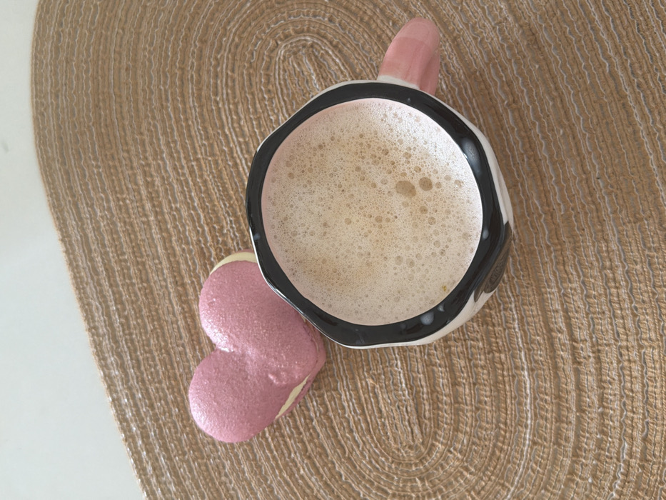 An overhead, static shot displays a heart-shaped macaron and a mug containing a frothy beverage, both resting on a light brown, woven placemat. The macaron, positioned in the upper-left, features a pink, textured top shell and an off-white filling. The mug, located in the lower-right, is white with irregular black markings resembling a cow print, a black rim, and a pink handle extending to the right. The liquid inside the mug is light brown, topped with a dense layer of foam. The placemat exhibits concentric oval weaving patterns and rests on a visible white surface in the upper-left corner. No individuals are present. The image provides no specific visual cues to identify its location within Tel-Aviv, Israel.
