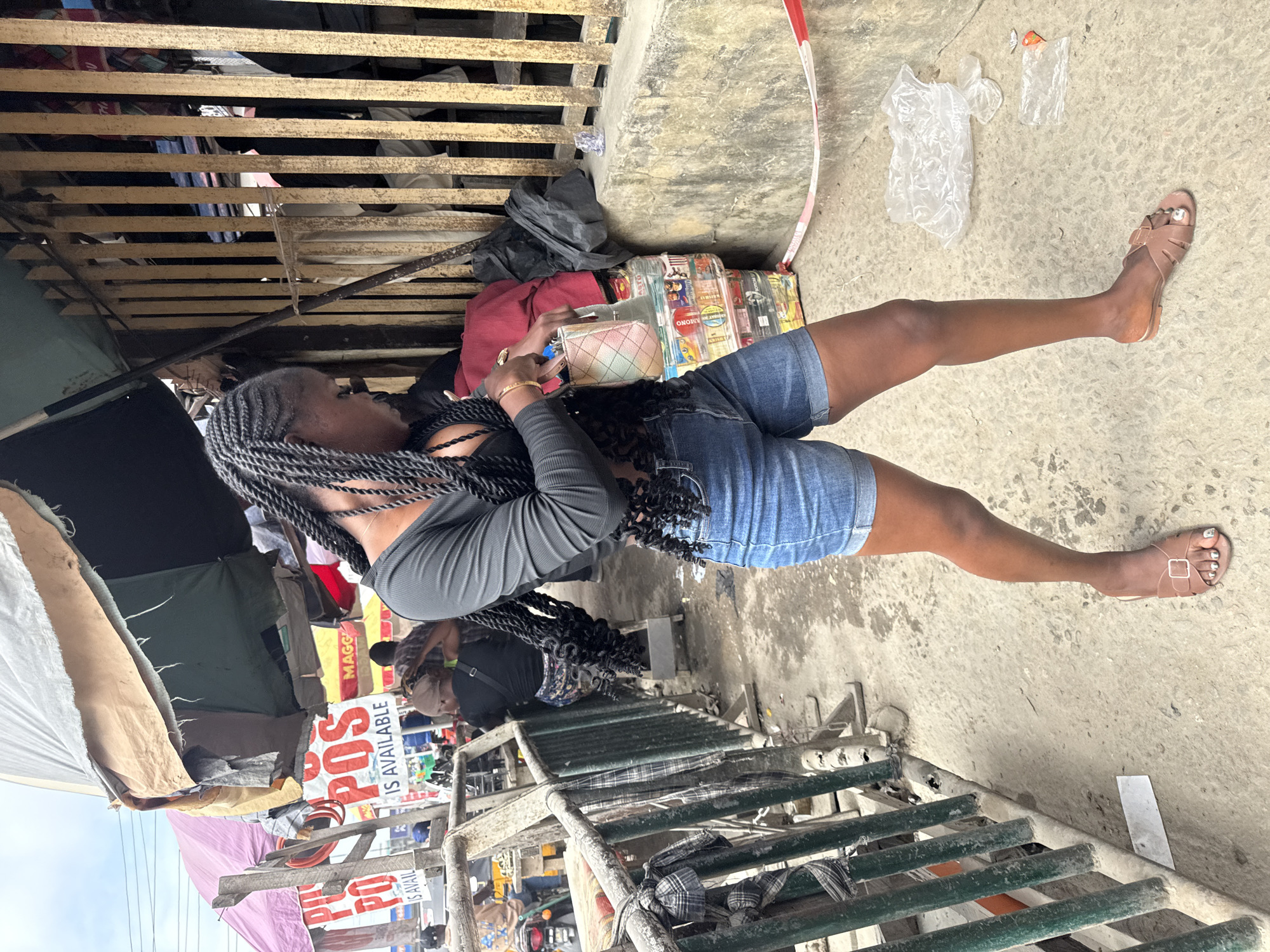 A Black woman with long, dark braided hair stands on a sidewalk in Lekki, Nigeria. She is wearing a black long-sleeved top with a cutout detail on the chest, denim shorts, and brown flat sandals. She is holding a small, multi-colored purse and her right hand is resting on top of it. Her left arm is bent at the elbow. Behind her, a stack of goods wrapped in plastic is visible, along with a yellow metal fence and a beige concrete wall. In the background, there are various market stalls with awnings and signs, including one that reads "POS IS AVAILABLE." Other people are visible in the background, some appearing to be vendors or customers. The ground is a paved sidewalk with some debris and plastic bags scattered around.