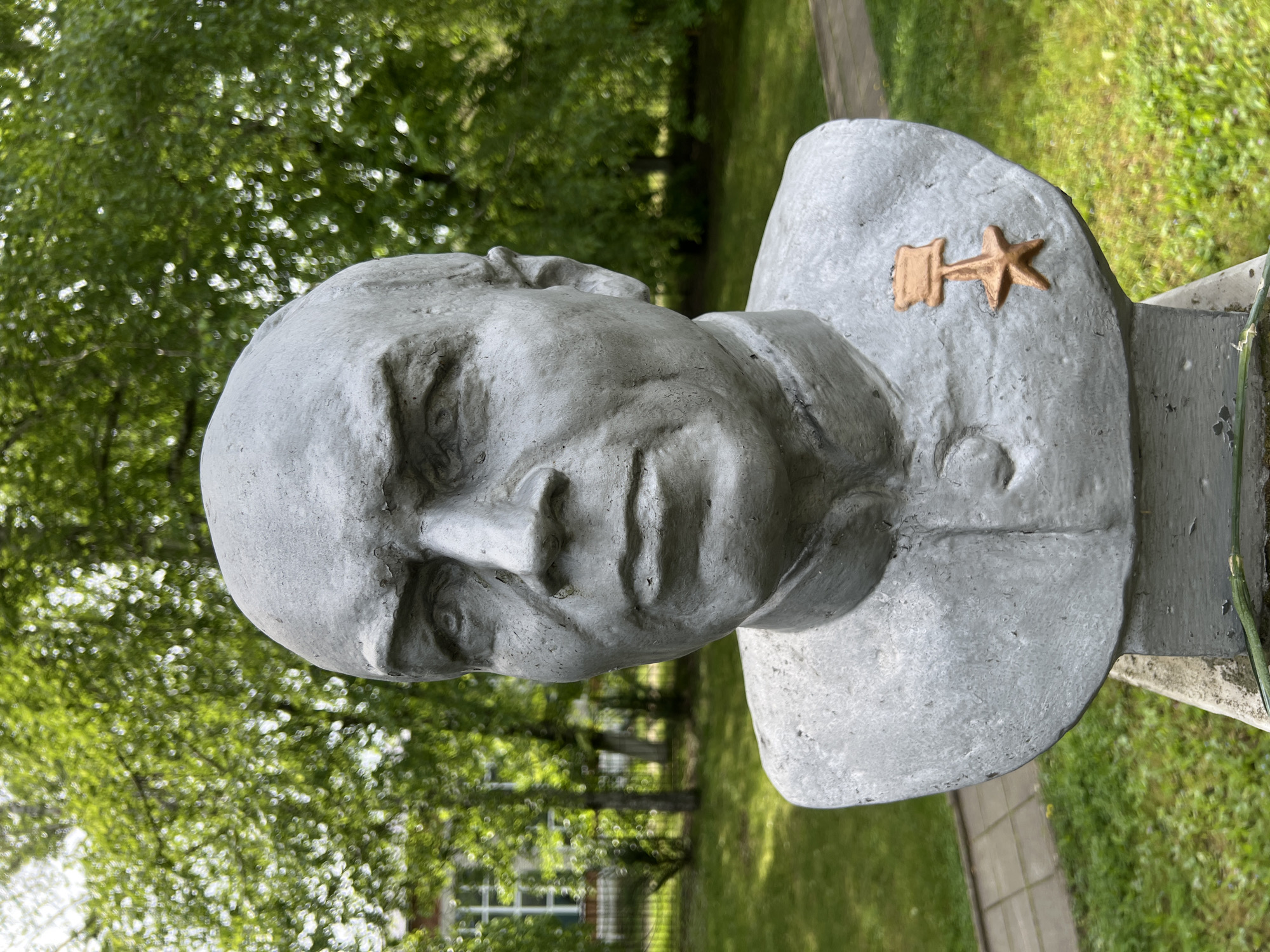 A bust of a man in military uniform is located outside in Goryachiy Klyuch, Russia. The bust is made of a grey material. The man is wearing a uniform with a high collar and a symbol that looks like a hammer and a star. The background includes green grass, trees, and a building.