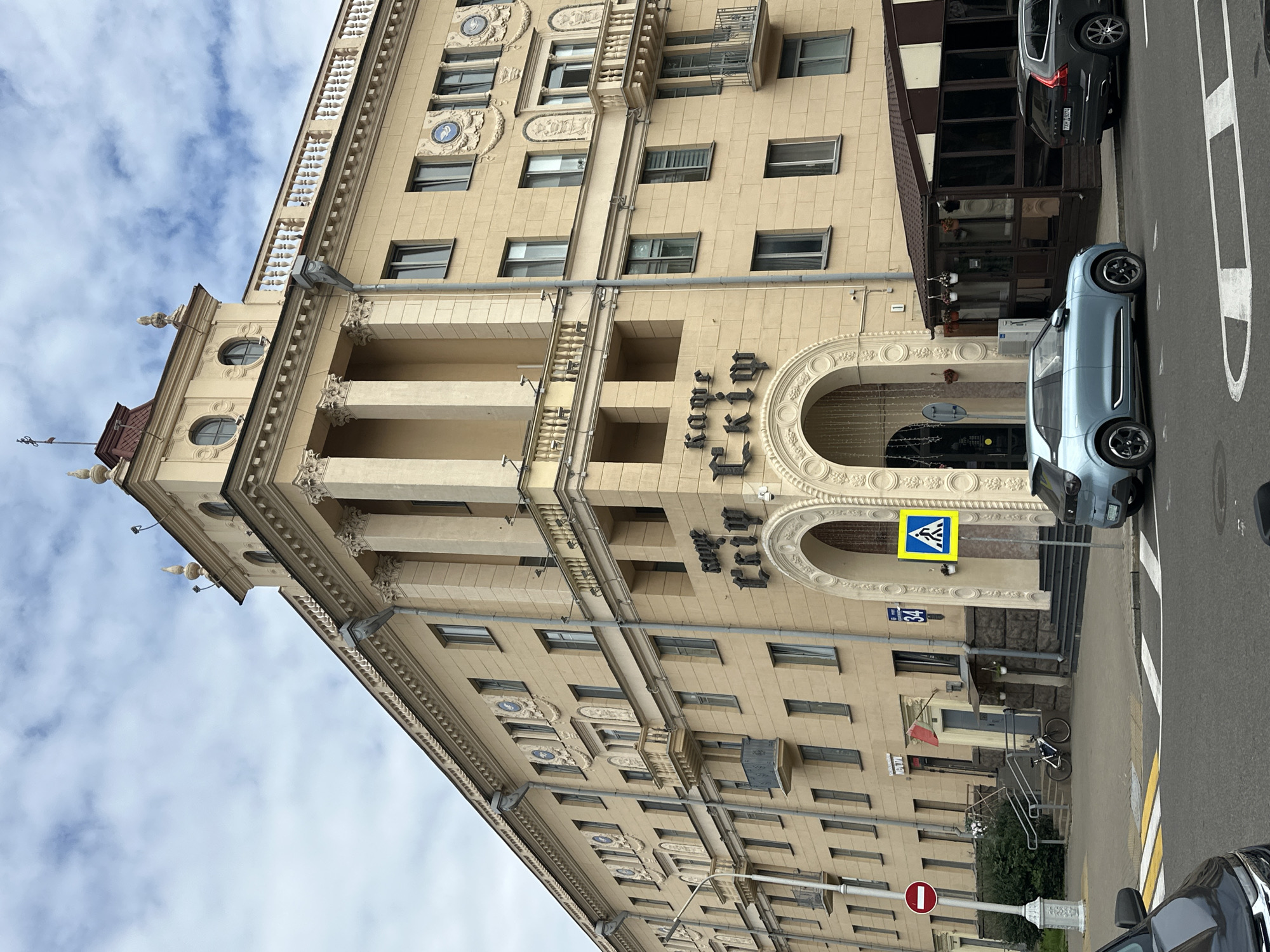 The image shows a building with a cafe named "Кафе Скiм" in Minsk, Belarus. The building is a multi-story structure with a classic architectural style, featuring columns, balconies, and decorative elements. The facade is painted in a light yellow color. In front of the building, there is a sidewalk with a pedestrian crossing and street signs. A light blue electric car is parked on the street, and a dark gray Volvo SUV is parked behind it. There are no people visible in the scene. The sky is cloudy, and the weather appears to be overcast.