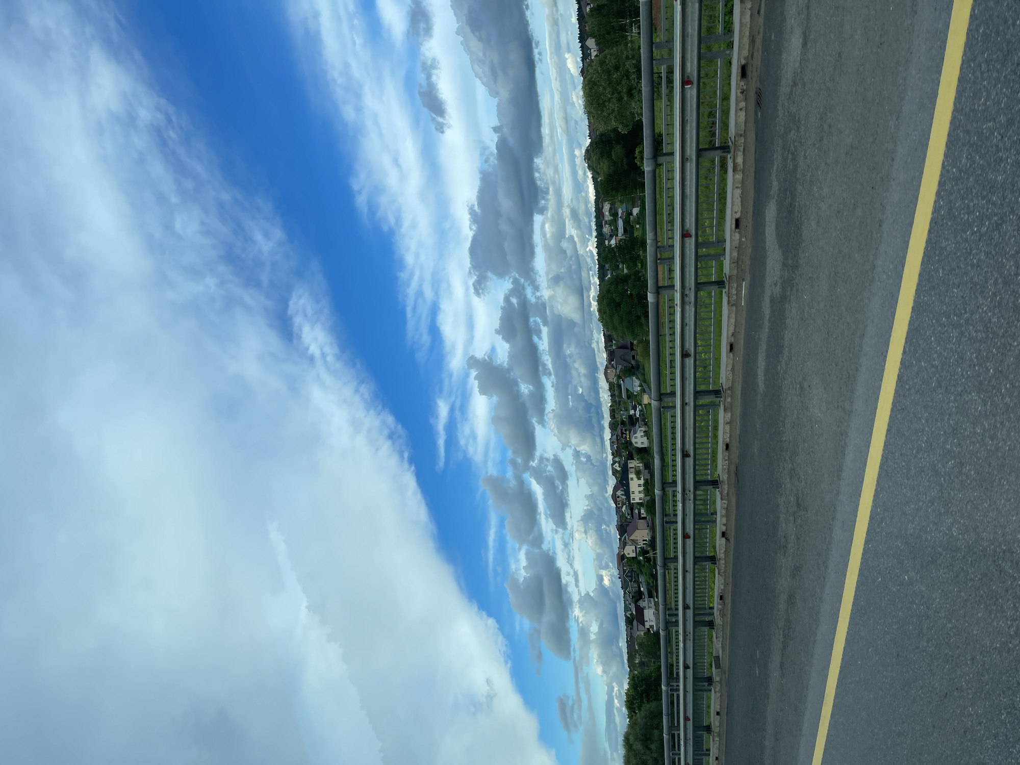 The image shows a roadside view from what appears to be a bridge or elevated roadway. A grey metal guardrail runs along the edge of the road. Beyond the guardrail, there is a line of buildings, including houses, partially obscured by trees. The sky above is blue with white and grey clouds. A yellow line marks the edge of the asphalt road. The picture's location is possibly near Бавыкино, Russia, due to the presence of buildings characteristic of Russian suburban or rural areas. The overall scene is calm, depicting a typical roadside view on a cloudy day.