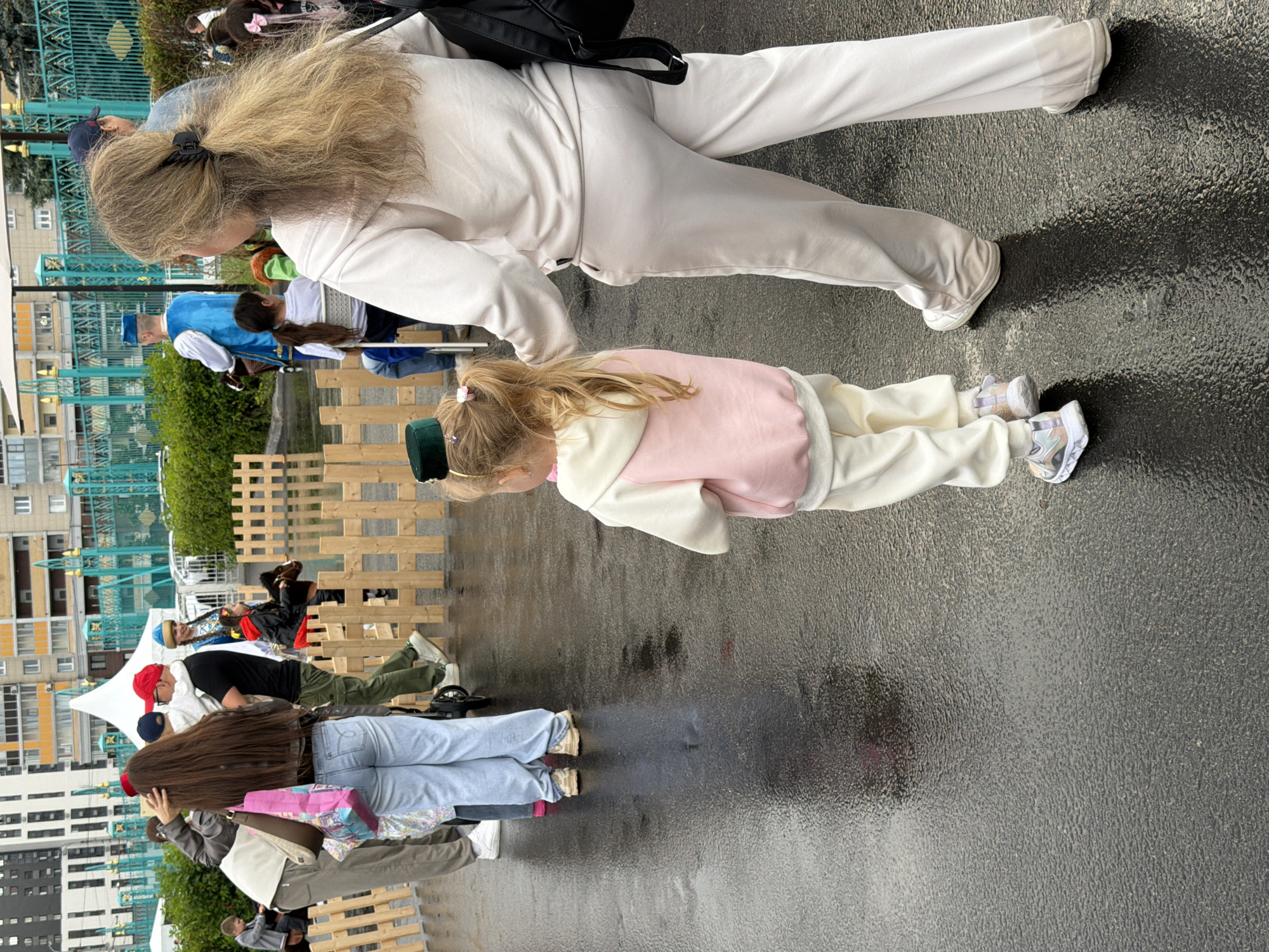An outdoor scene shows a wet asphalt ground with reflections of people and a white tent. In the foreground, a woman with long blond hair and a black hair clip is seen from behind, wearing a cream-colored tracksuit. Beside her, a young girl with blond pigtails, also wearing a cream and pink tracksuit and a green hat, walks away from the camera. A wooden fence is visible behind them, separating them from other people and a large white tent. In the background, several buildings and a teal-colored fence can be seen. A man in a blue and white historical costume stands near the fence, and other people are present in the background, some of whom appear to be children interacting with a ride. The overall atmosphere suggests an outdoor event or fair.