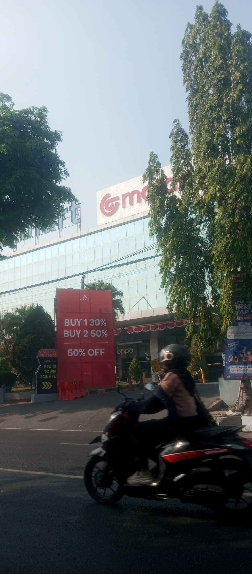 This is a daytime, eye-level outdoor photograph in the city of Kediri, Indonesia. The image captures a street scene with a modern building in the background and a person riding a motorcycle in the foreground.
The building, likely a shopping center or department store, features a large, glass facade. Prominently displayed on the building's exterior are large signs. One sign, in red, advertises a sale with text that reads "BUY 1 30%", "BUY 2 50%", and "50% OFF". Above this, the logo and name "Gm" (likely part of a larger brand name) are visible in red and white. To the left of "Gm", the letters "rt" are visible in silver. To the right of the "Gm" sign, a green tree with dense foliage is visible. Another sign for "oppo" is visible on the building's lower level.
In the foreground, a person wearing a helmet and dark clothing is riding a dark-colored motorcycle. The motorcycle is in motion, creating a slight blur in the image. The person appears to be looking forward. The road surface is asphalt, with a white line marking the lane. Trees and a blue sky with some haze are visible in the background. To the left of the red sale banner, a directional sign for "PARKING ENTRANCE" and "KEDIRI TOWN SQUARE" is visible.