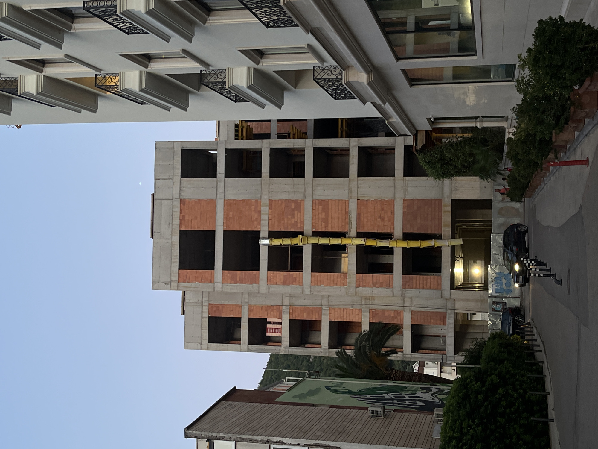 This image captures a view of a street in Budva, Montenegro, with a partially constructed building dominating the central frame. The building's concrete frame and brickwork are visible, with empty window openings. A yellow chute extends down the side of the structure. To the right, a completed building with balconies and windows is present. On the left side of the street, a building with a mural is partially visible, along with lush green foliage and a palm tree. Two cars are parked on the street, one of which is a black SUV. The sky is clear and blue, suggesting it is daytime. The scene appears to be a street in a residential or commercial area undergoing development.