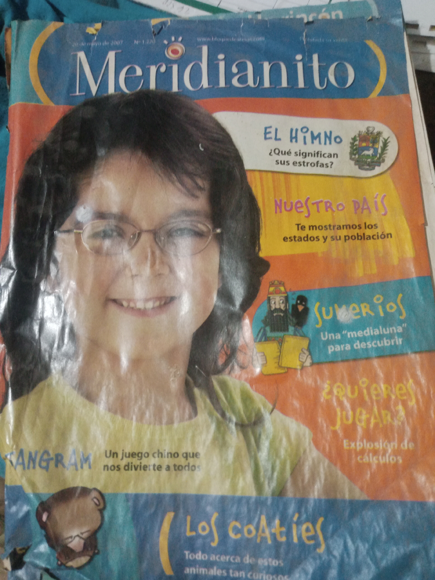 This is a close-up, angled shot of the cover of a Venezuelan children's magazine titled "Meridianito." The cover features a smiling young girl with glasses wearing a yellow shirt, her face taking up a significant portion of the frame. Various articles and illustrations are visible on the cover, including text related to "EL HIMNO," "NUESTRO PAÍS," "SUMERIOS," "TANGRAM," and "LOS COATÍES." The magazine appears to be from May 2007, with a publication number of "N° 1.370." There is no discernible information within the image that indicates a specific location in the city of Santa Ana, Venezuela. The image is taken from above, with the magazine cover laid flat, showing signs of wear and some crinkling.