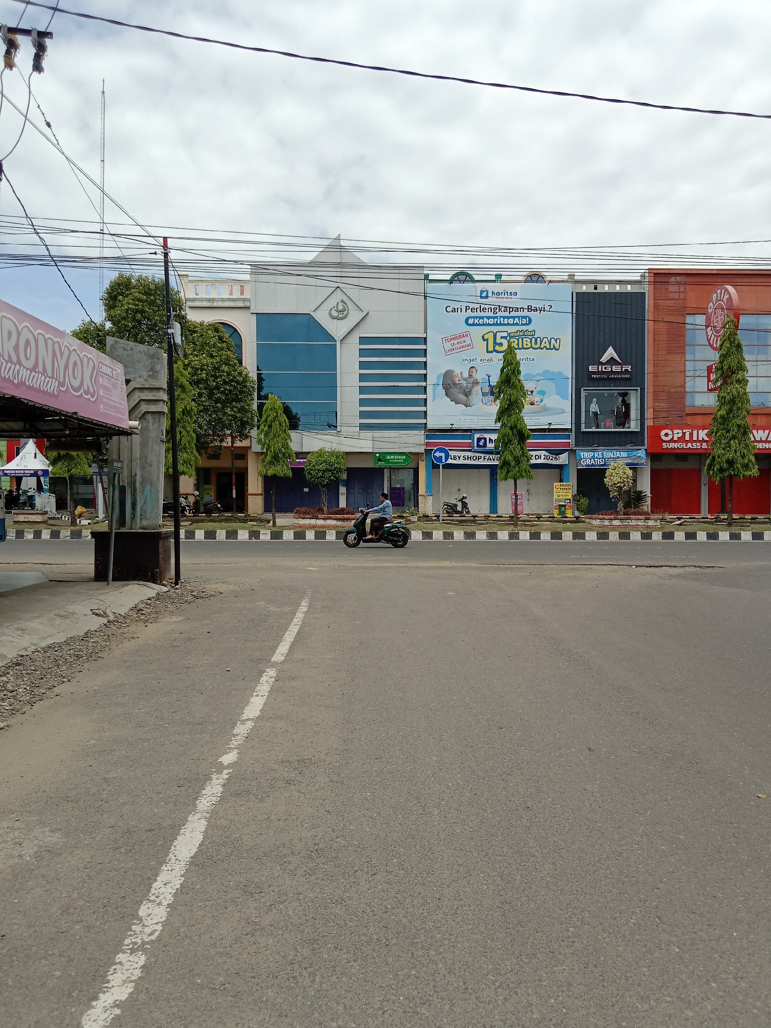 The image depicts a street scene in Lhokseumawe, Indonesia, with a row of commercial buildings lining the opposite side of the road. A scooter with a single rider is traveling on the road, heading right. The buildings exhibit different facades and signage, including "Haritsa baby shop" (Cari Perlengkapan Bayi), "Eiger Tropical Adventure," and "Optik." Trees are planted along the sidewalk in front of the buildings. A road marking is visible in the foreground, delineating a traffic lane. The sky is overcast. Power lines crisscross overhead. A sign that says "RONYOK rasmanan" on the left.