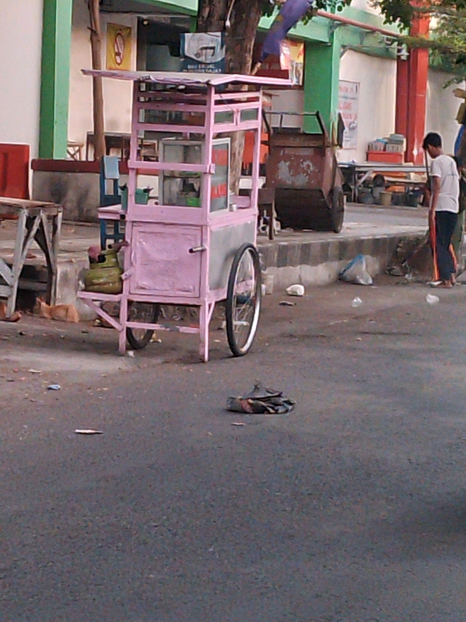 The image depicts a street scene in Kertosono, Indonesia. A pink food cart with a gas tank is parked on the right side of the frame. A person is sweeping the street in front of a large, rusty garbage container. The ground is covered with litter, including plastic bags and debris. A wooden table and chairs are visible in the background, suggesting a seating area. A tree with a flag is also present.
