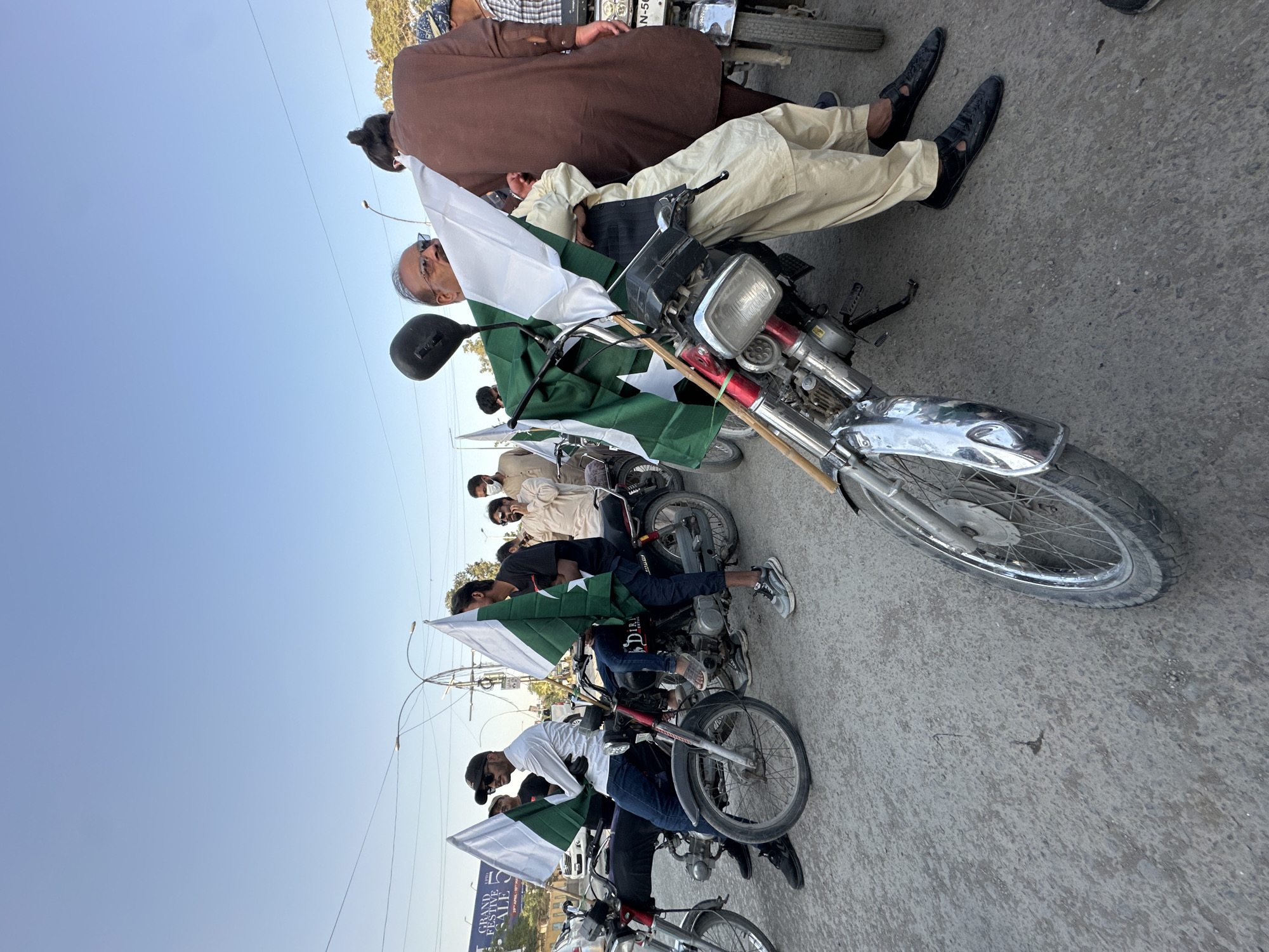 A street in Quetta City Tehsil, Pakistan, is filled with a procession of people on motorcycles, likely celebrating a patriotic event. The riders are carrying Pakistani flags, green with a white crescent moon and star, and some have white flags as well. Many individuals are wearing traditional Pakistani clothing, such as shalwar kameez. The motorcycles are standard models, and the street is paved. Overhead, electrical wires crisscross the sky.