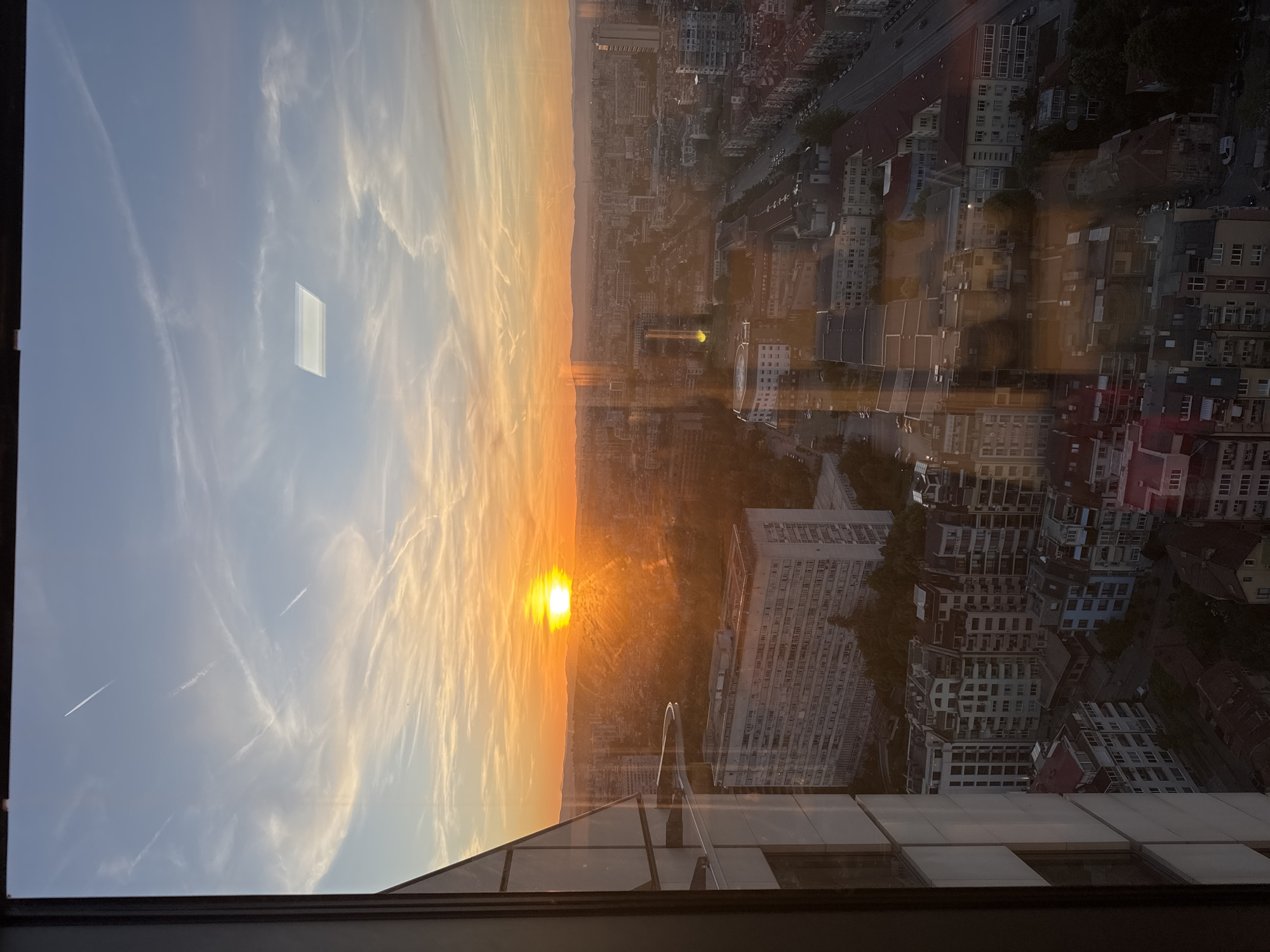 The image shows a sunset view over Sofia, Bulgaria, likely seen from a high-rise building. The sun is setting on the horizon, casting an orange glow across the sky with streaky clouds. Below, a cityscape is visible, showing a mix of building types including apartment blocks, offices, and residential structures. Roads and vehicles can also be seen. There is a clear reflection in the glass pane from which the image is taken. A rectangle-like shape can be seen reflecting in the window. No people are visible in the image.