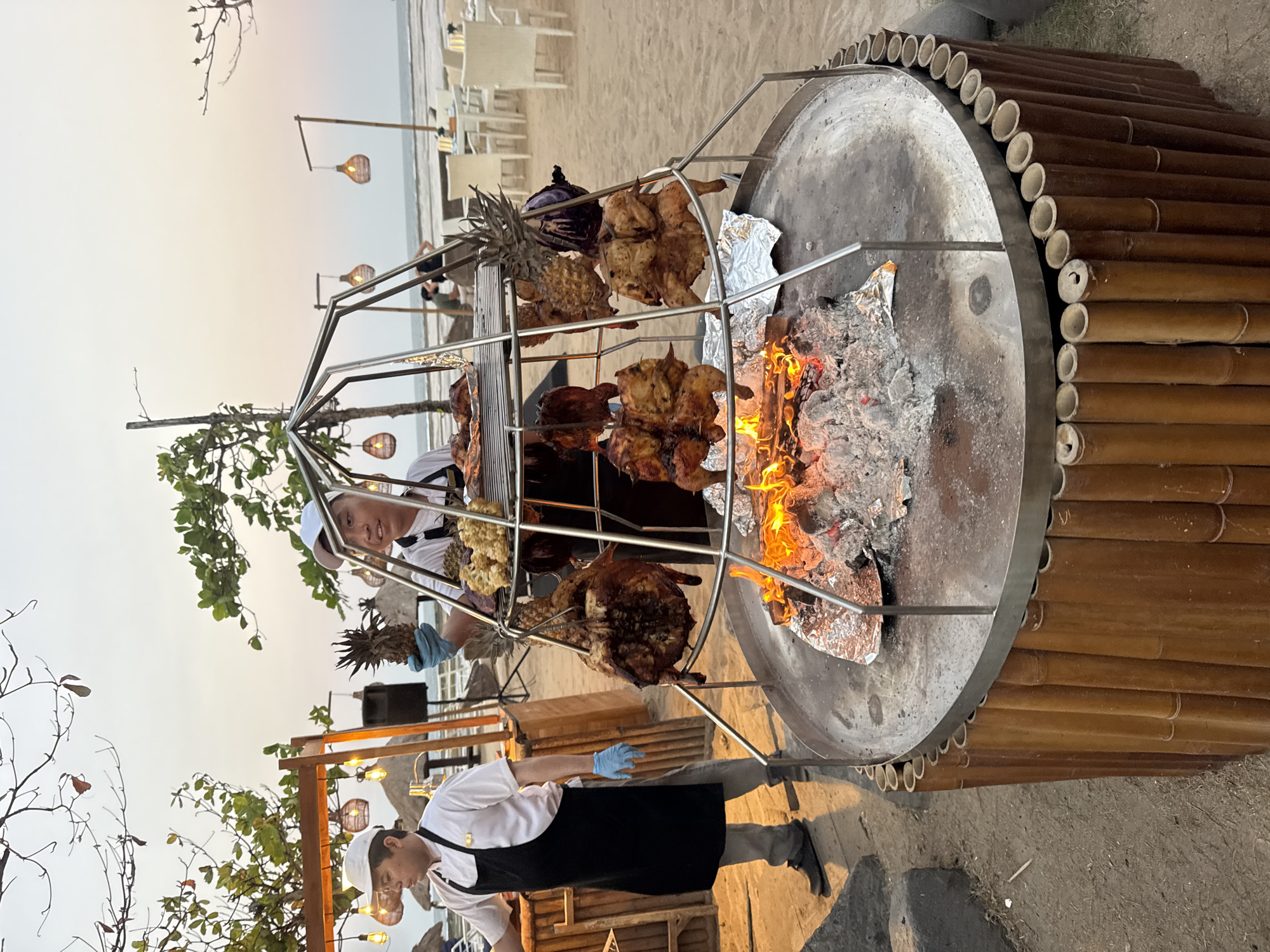 This image depicts an outdoor barbecue setup on a sandy beach in Kuta Selatan, Indonesia. Two individuals, likely chefs, are attending to a large rotisserie grill. One chef, wearing a white chef's hat, white shirt, black apron, and blue gloves, stands to the left, observing the grill. The second chef, also in a white chef's hat and white shirt with a black apron, is on the right and is holding a pineapple. The rotisserie grill is circular with a metal top and a bamboo-wrapped base, containing a large fire and glowing embers. Several whole chickens and pineapples are suspended from the metal frame of the rotisserie, being cooked over the open flame. In the background, beach chairs and tables are visible, along with thatched umbrellas, suggesting a dining area. The sky is overcast with the soft light of dusk. The scene suggests a culinary event taking place at a beachside restaurant or resort.